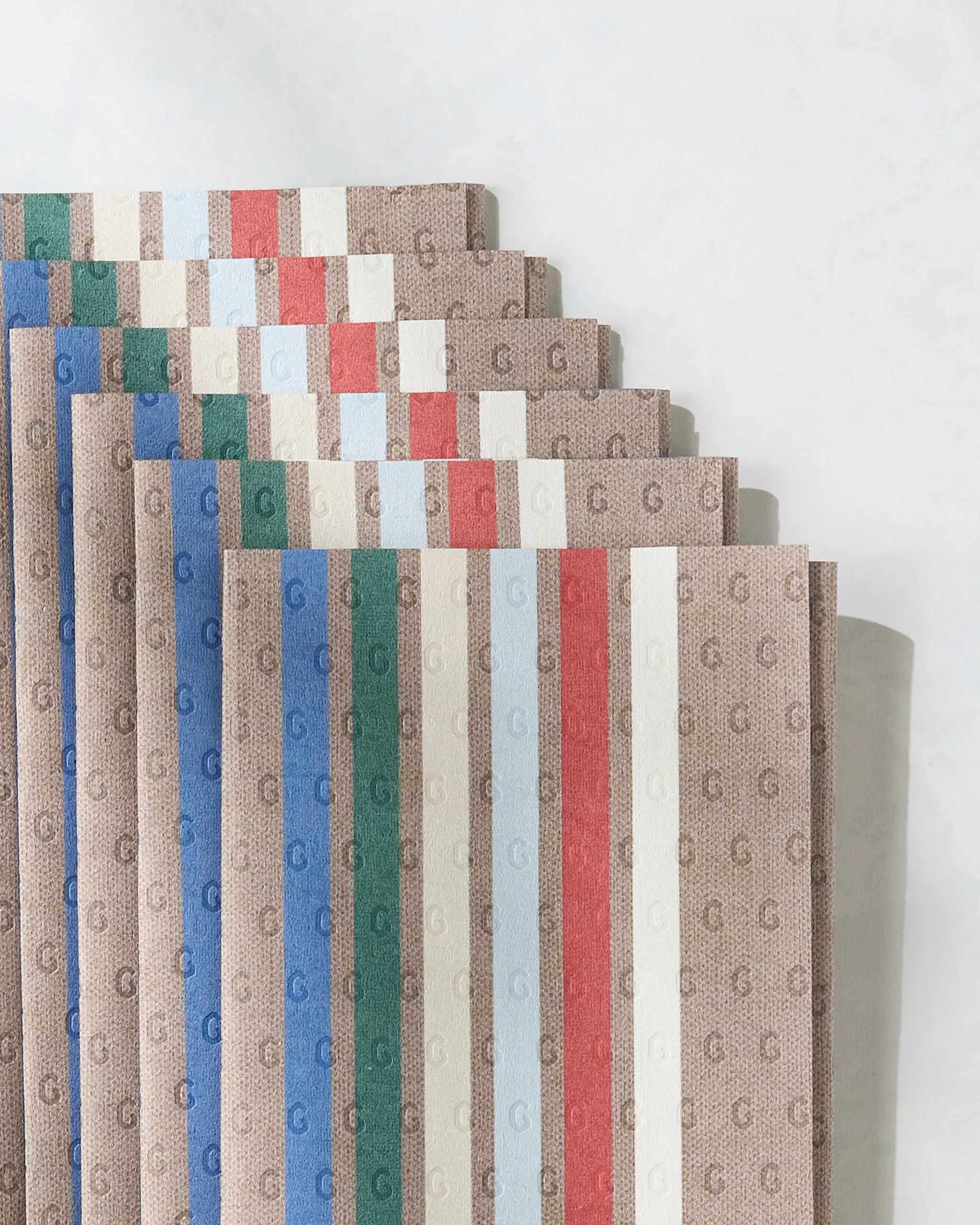 Winterhaus by Geometry—folded fabric pieces with a tan background, repeating G monogram, and vertical blue, green, red, and white stripes. An eco-friendly Not Paper Towels alternative on a light surface.