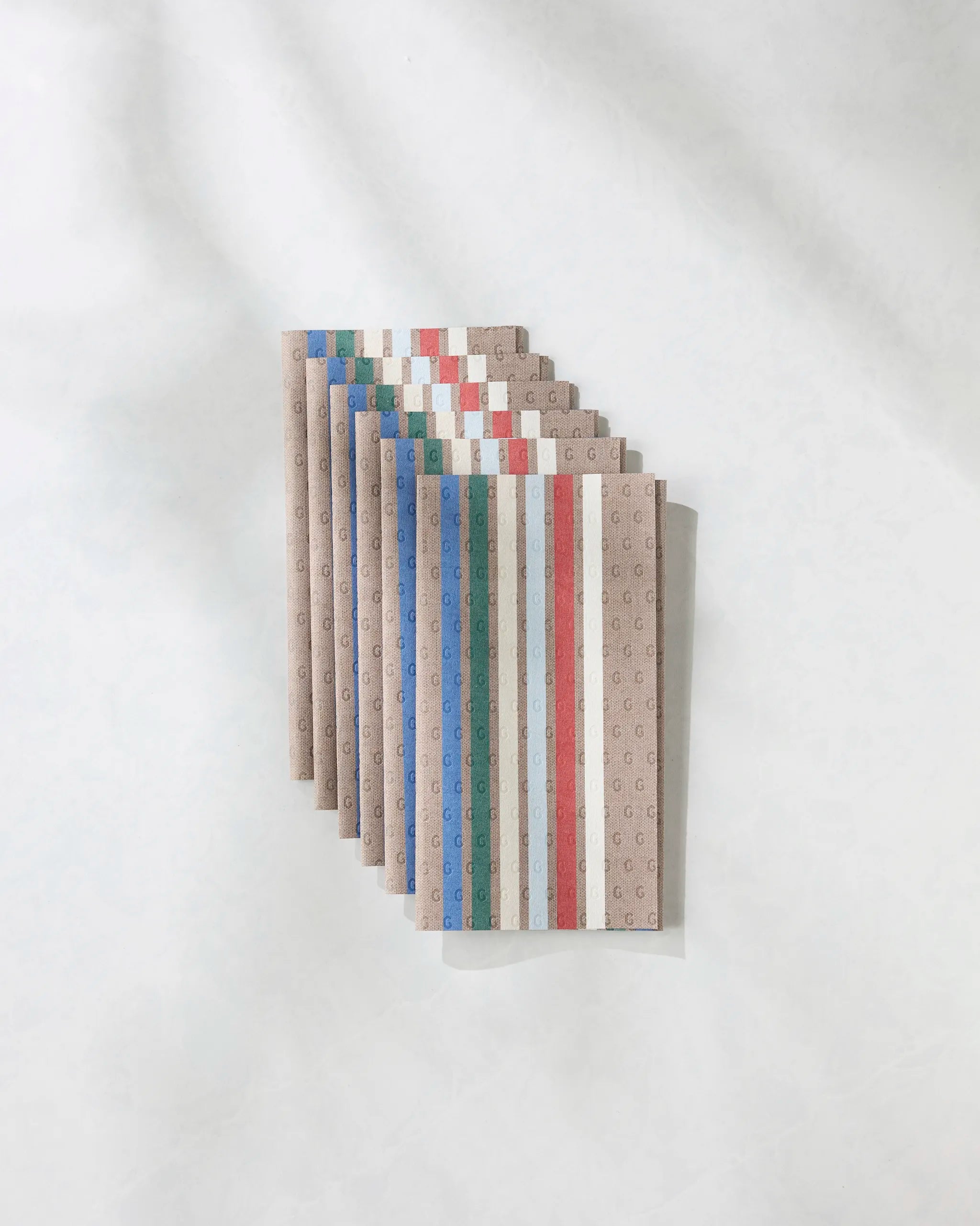 Nine Winterhaus notebooks by Geometry, featuring brown covers with vertical blue, green, red, and white stripes and subtle G patterns, are neatly stacked on a light textured surface—an eco-friendly alternative to Not Paper Towels.