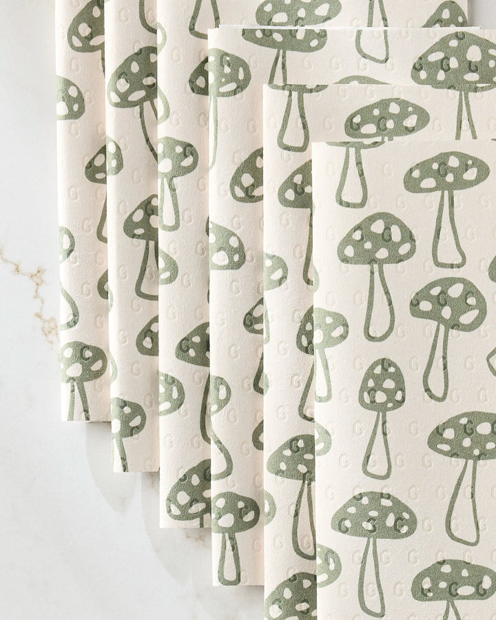 Several Woodland Mushroom napkins by Kayla Ann, made from recycled paper towels and featuring a repeating green mushroom with white spots design, are folded in a fan shape on a white surface.