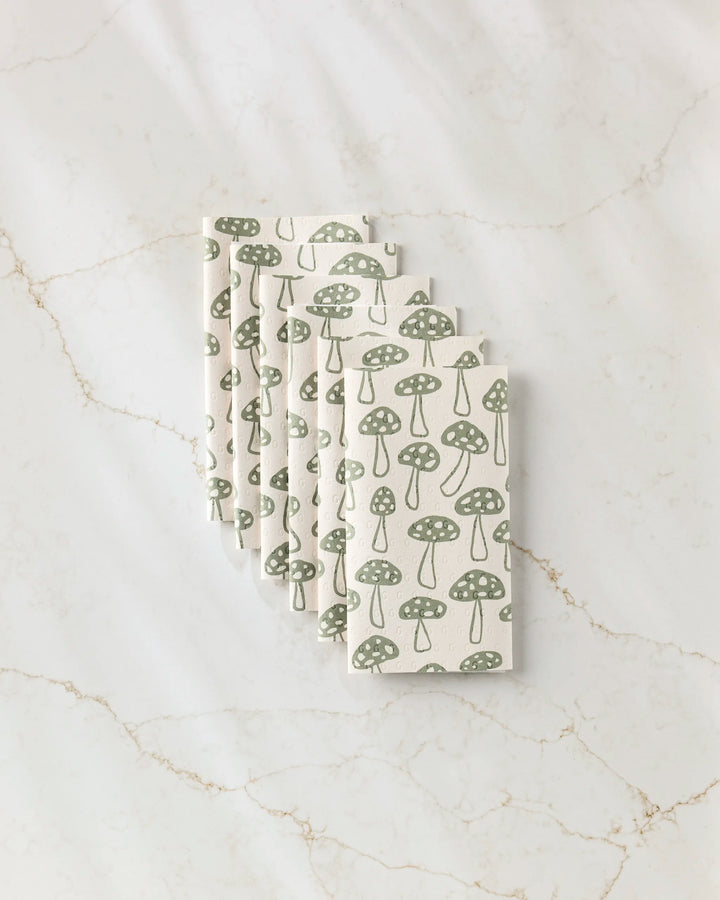 Six folded Kayla Ann Woodland Mushroom white cloth napkins with a green mushroom design, perfect as eco-friendly paper towel alternatives, are neatly arranged on a white marble surface with tan veining.