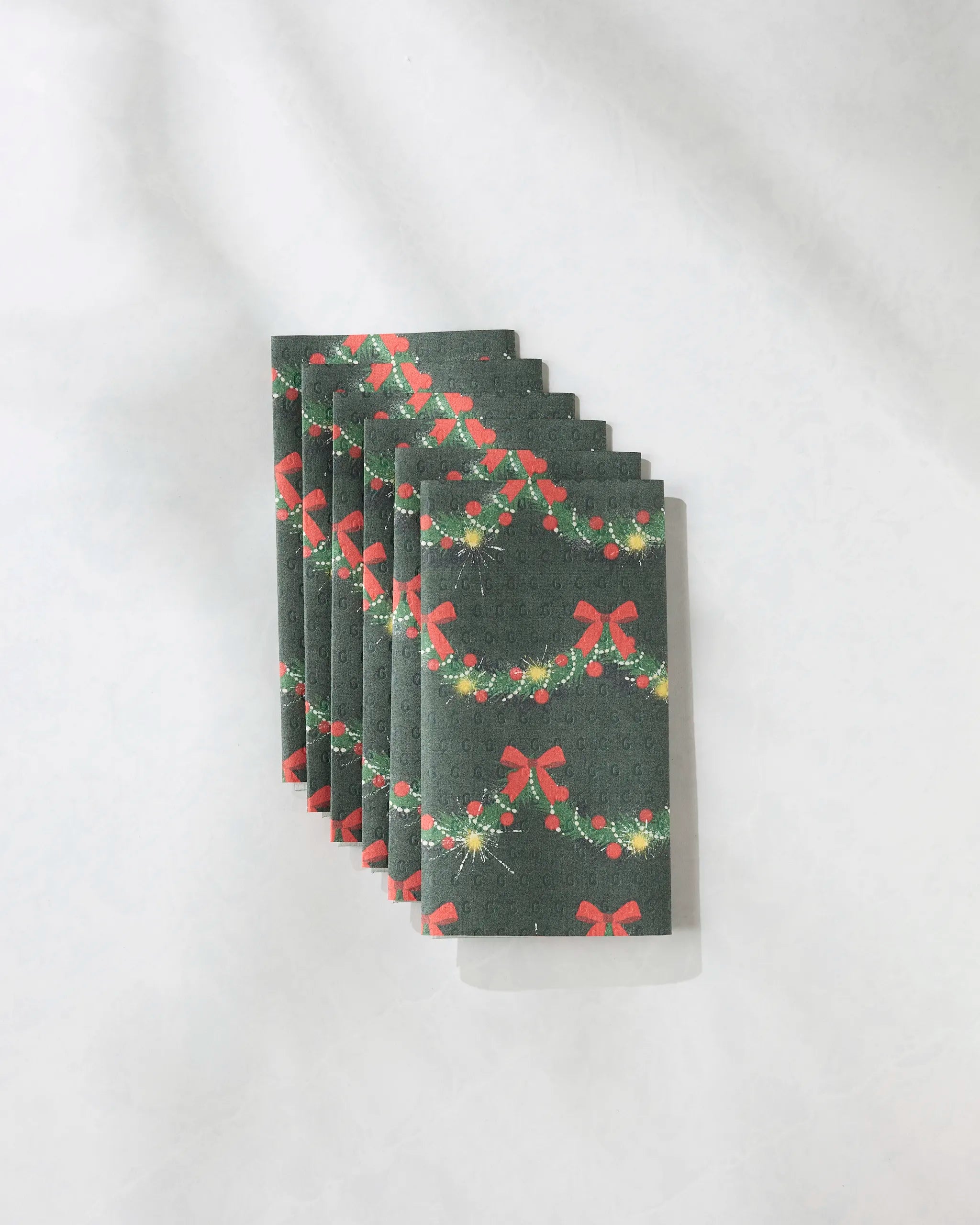 Six folded Yuletide Garland reusable towels by Geometry, dark green with red bows, green garlands, and ornaments, fanned out on a light background—sustainable, festive alternatives to paper towels for the holidays.