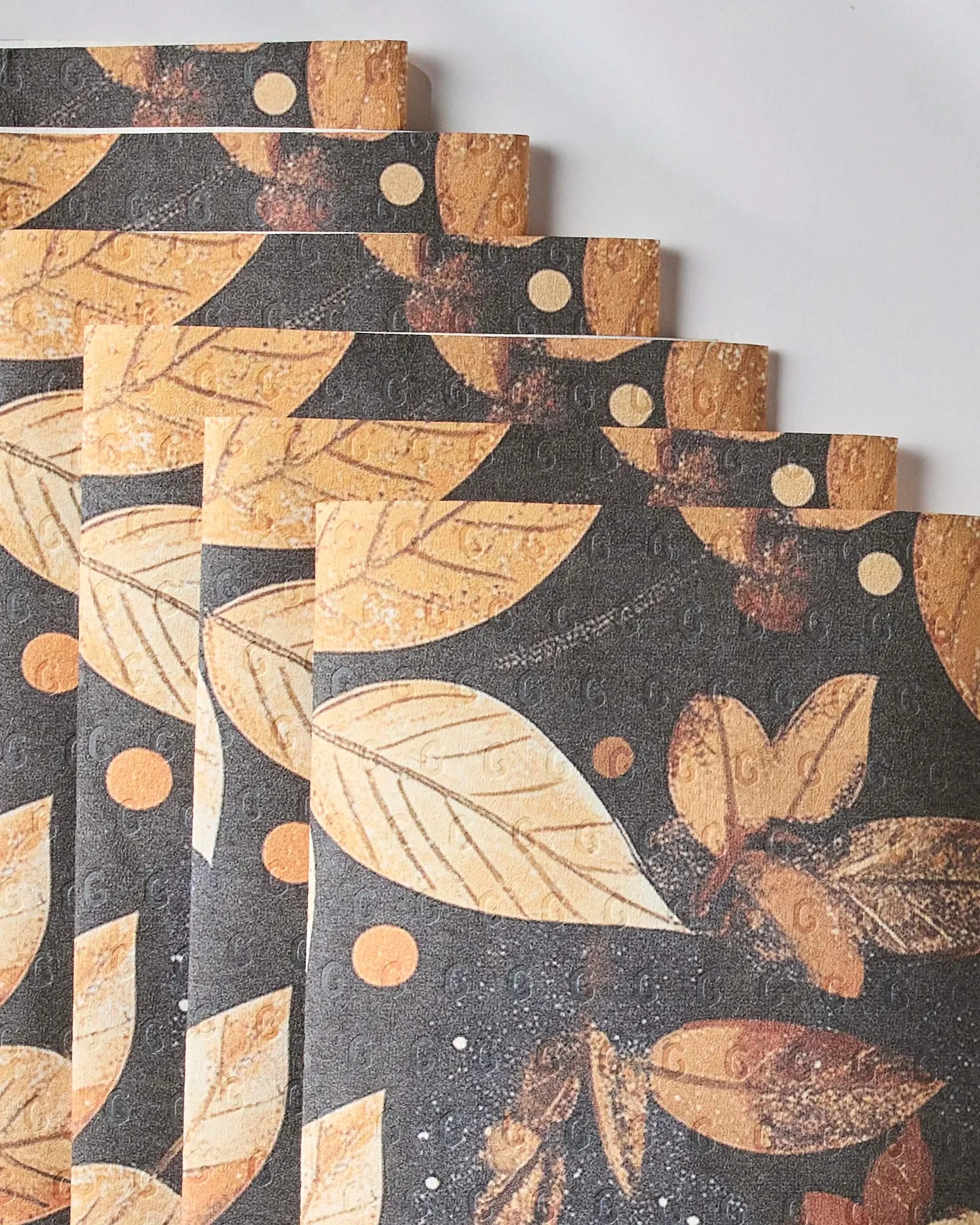 Five sheets of Geometrys Botanical Wonderland reusable paper towels, featuring a yellow and orange leaf and dot pattern on a dark background, are fanned out over a white surface.