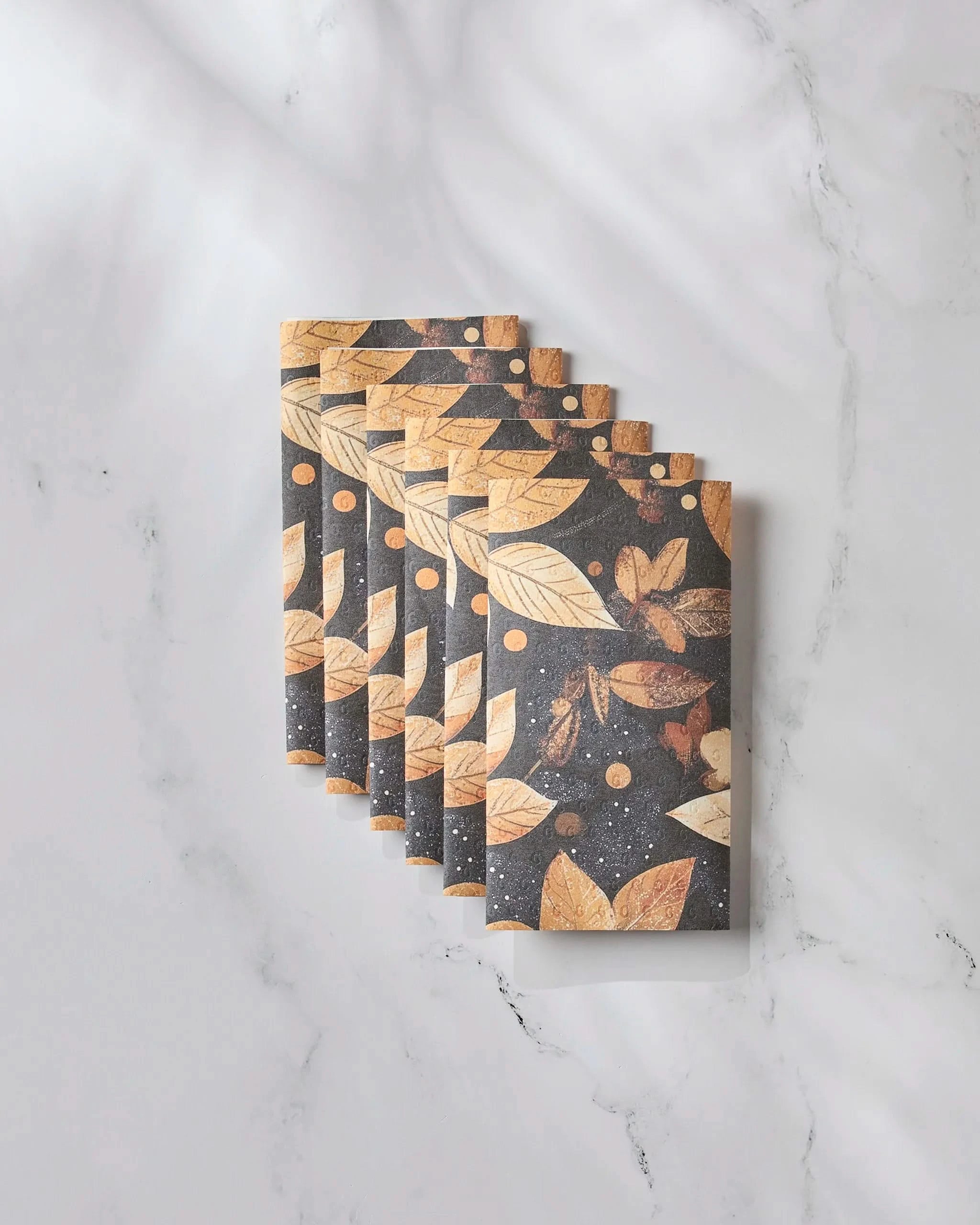 Five identical Botanical Wonderland notebooks by Geometry, featuring a brown, gold, and black leaf pattern, are neatly arranged in an overlapping row on white marble, offering an elegant look reminiscent of eco-friendly kitchen towels.