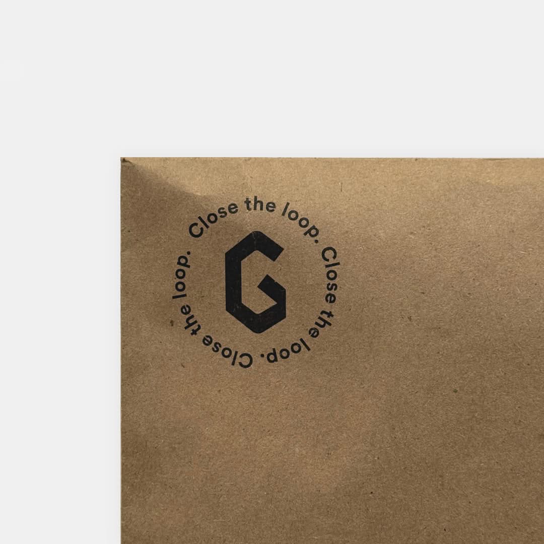 The GEOMETRY Geometry Take Back Bag is a brown paper envelope featuring a black circular Close the loop. stamp around a bold G, displayed on a white background.