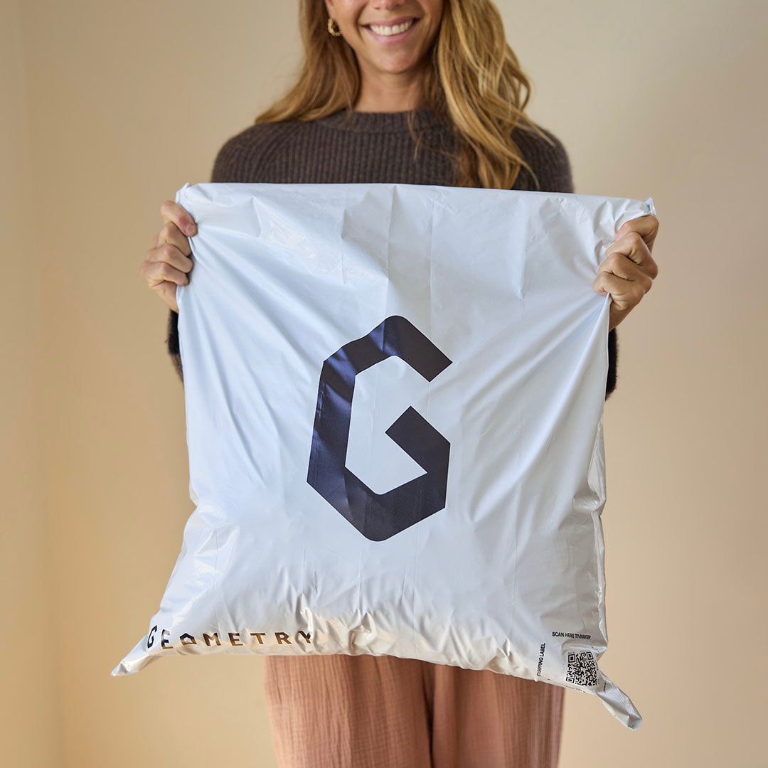 A woman with long hair smiles while holding a full Geometry Take Back Bag by GEOMETRY, featuring a bold black G and the brand name printed at the bottom.