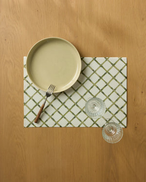 A beige plate with a fork sits on a white and green checkered Geometry Bamboo Lattice placemat, next to two empty wine glasses on a wooden table, as seen from above.