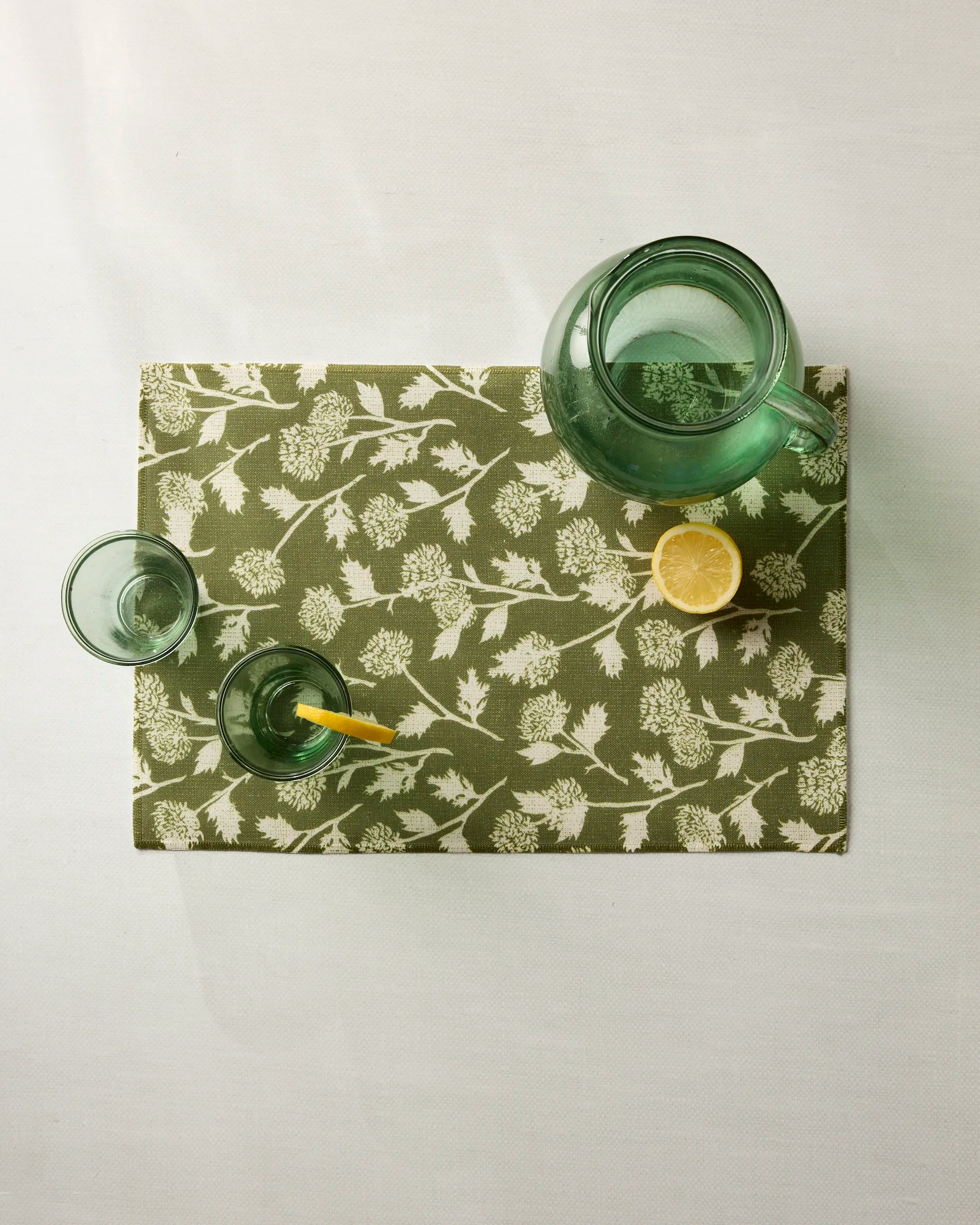 A green glass pitcher and two glasses, one with a yellow straw and lemon slice, sit atop the Geometry Bloom Thistle Green placemat beside a half lemon on a crisp white surface.