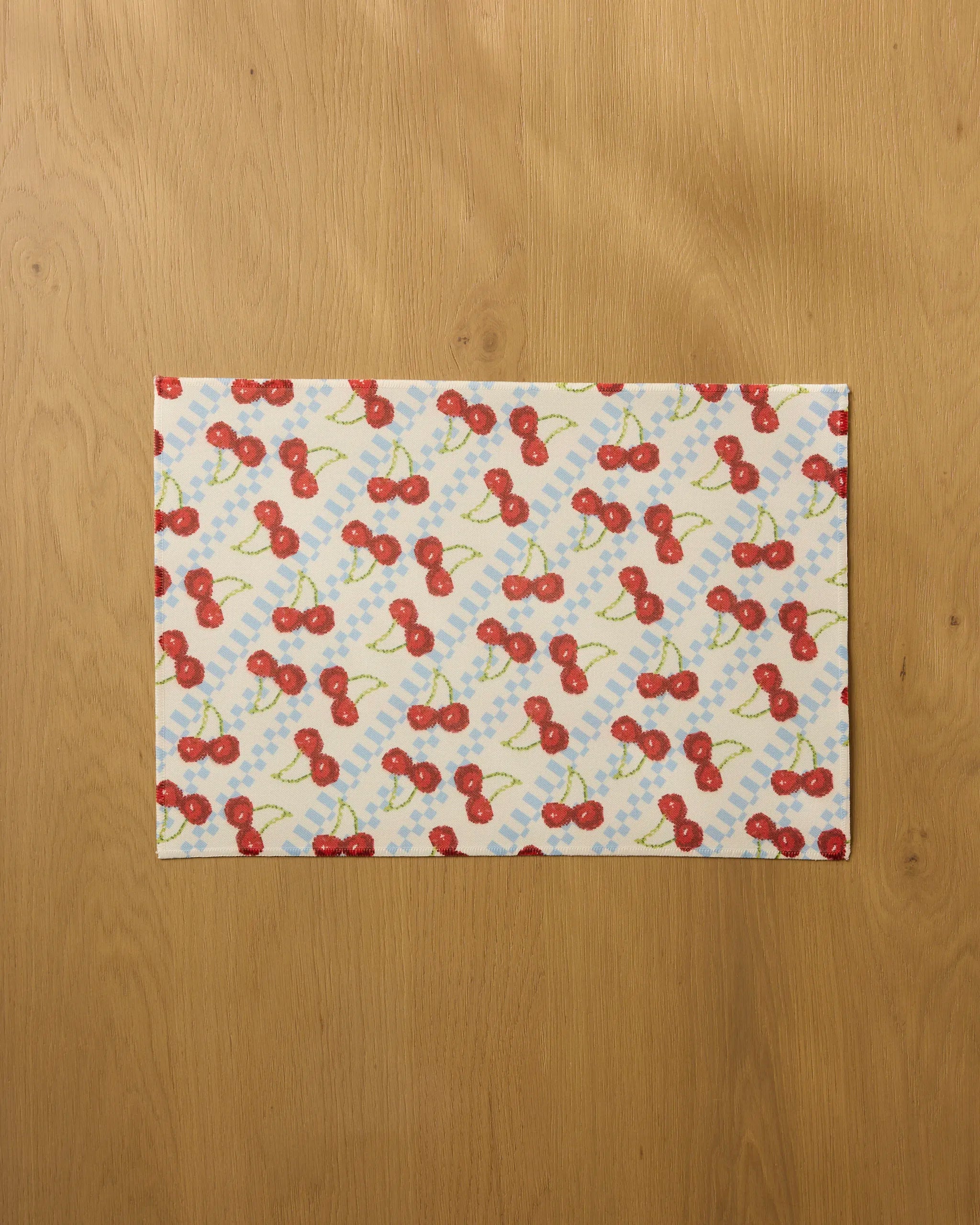 The Geometry "Cherry Pop" placemat features a diagonal pattern of red cherries, green stems, and blue dots on white. Rectangular and machine washable, it is shown on a light wooden surface.