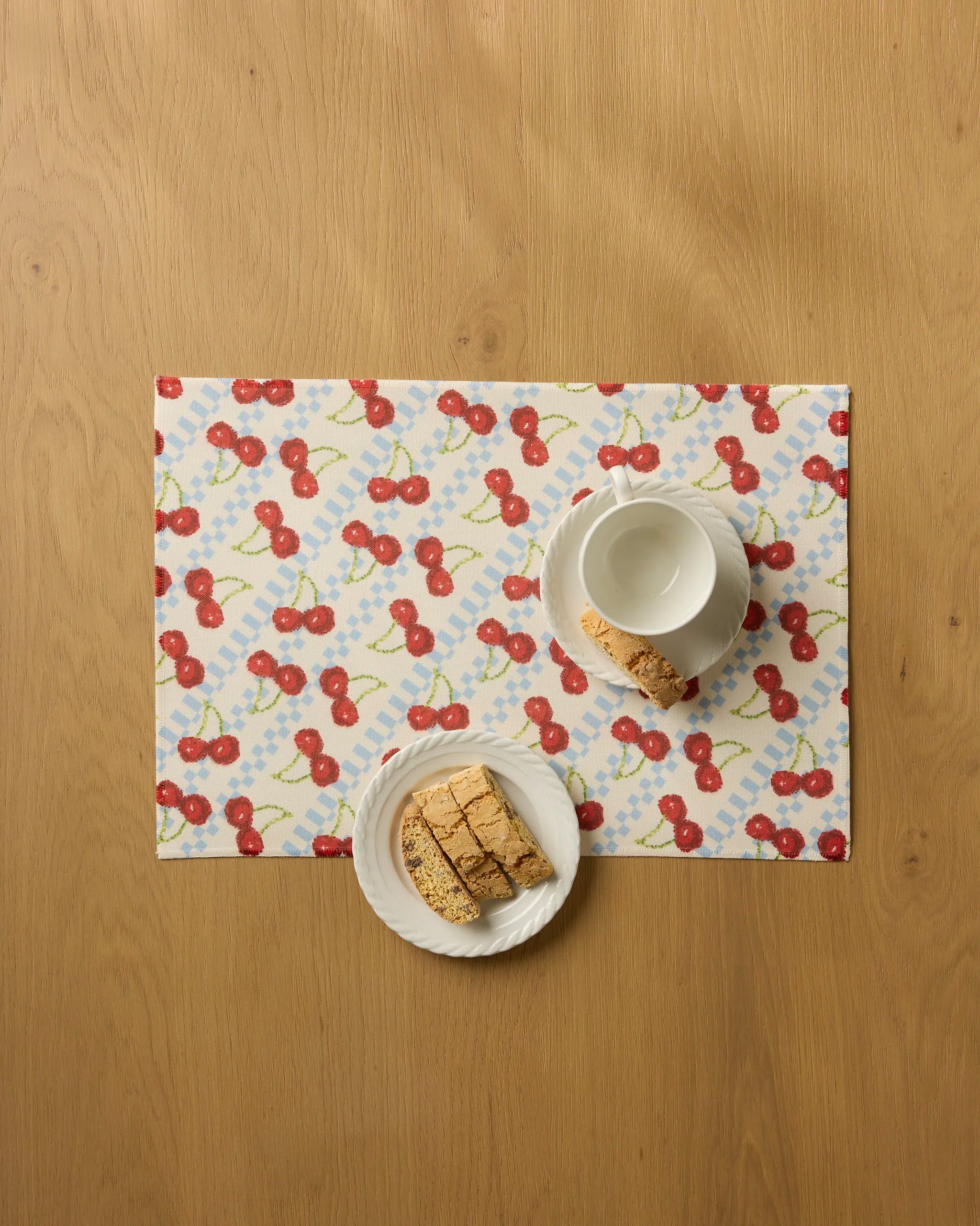 A Geometry Cherry Pop placemat with cherry patterns holds a white cup and saucer next to a plate of sliced biscotti, all arranged on a light wooden table.