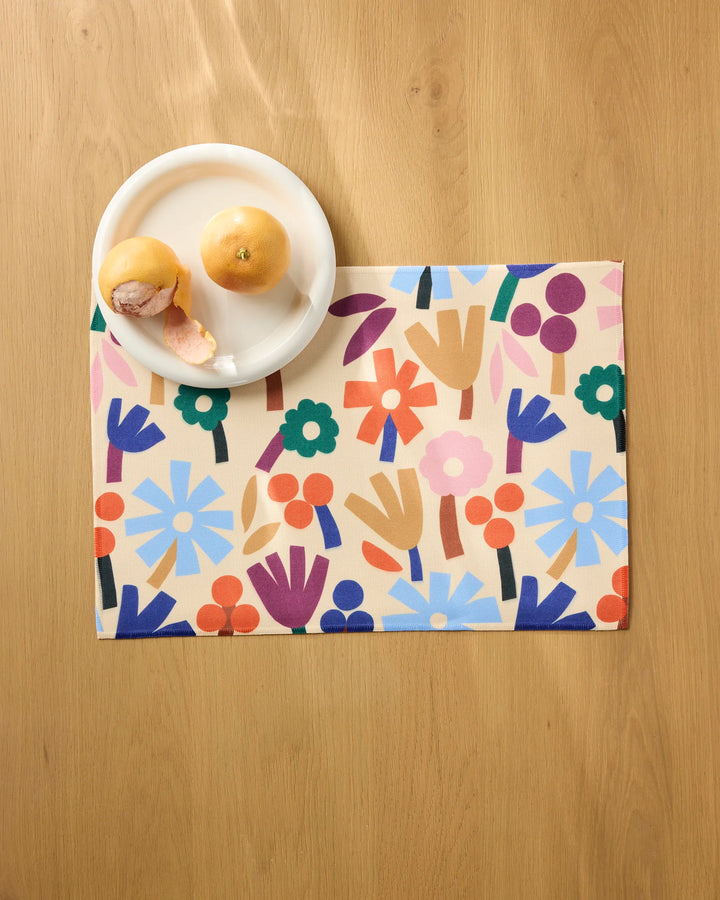A white plate with two oranges, one partially peeled, sits on a light wooden table beside vibrant Cool Flowers placemats by Jennifer Bouron, featuring abstract floral patterns in blue, red, green, orange, and purple.
