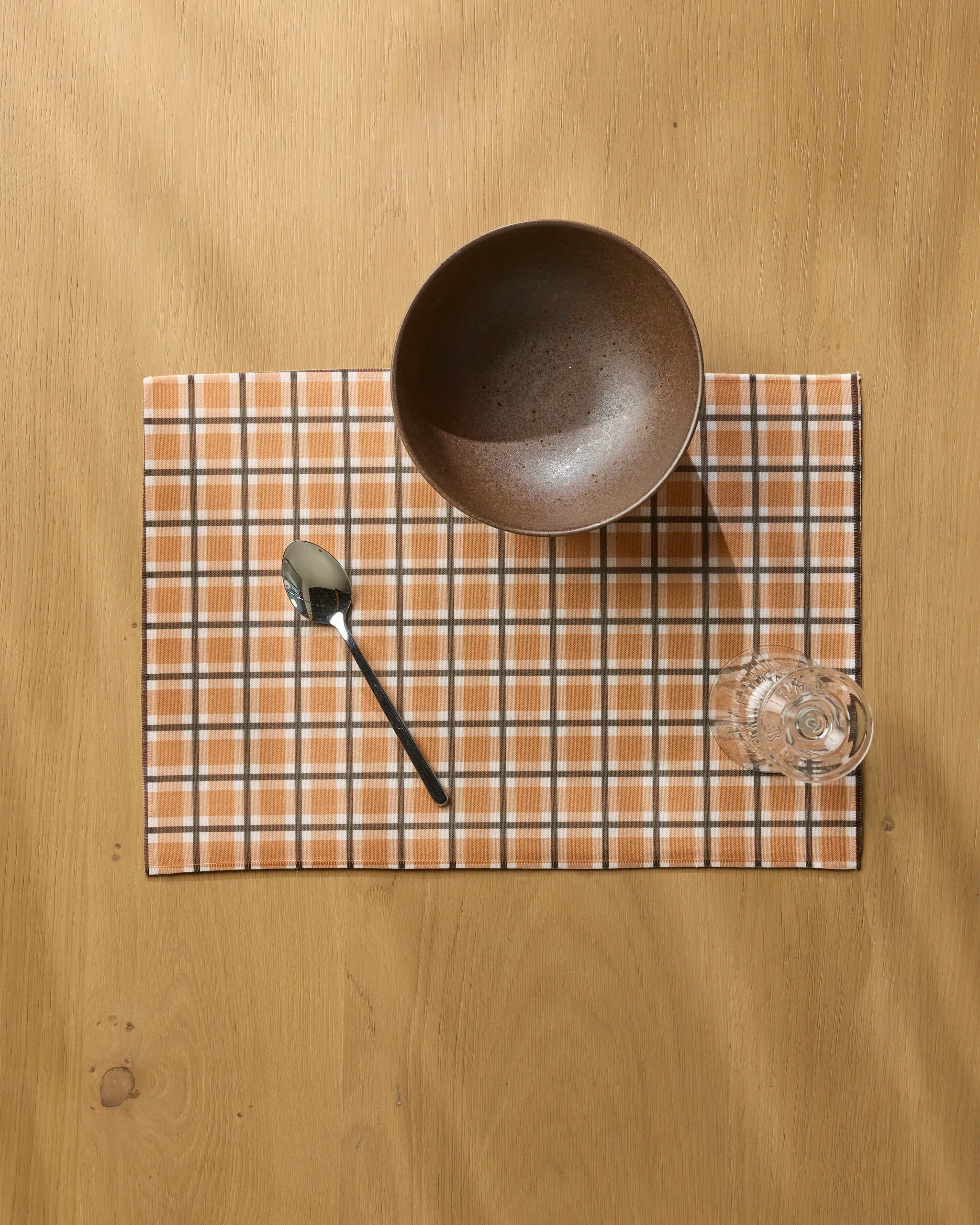 A brown bowl, metal spoon, and clear glass rest on a plaid orange and white Country Pantry placemat by Geometry atop a light wooden table—ideal for displaying your favorite machine washable placemats.