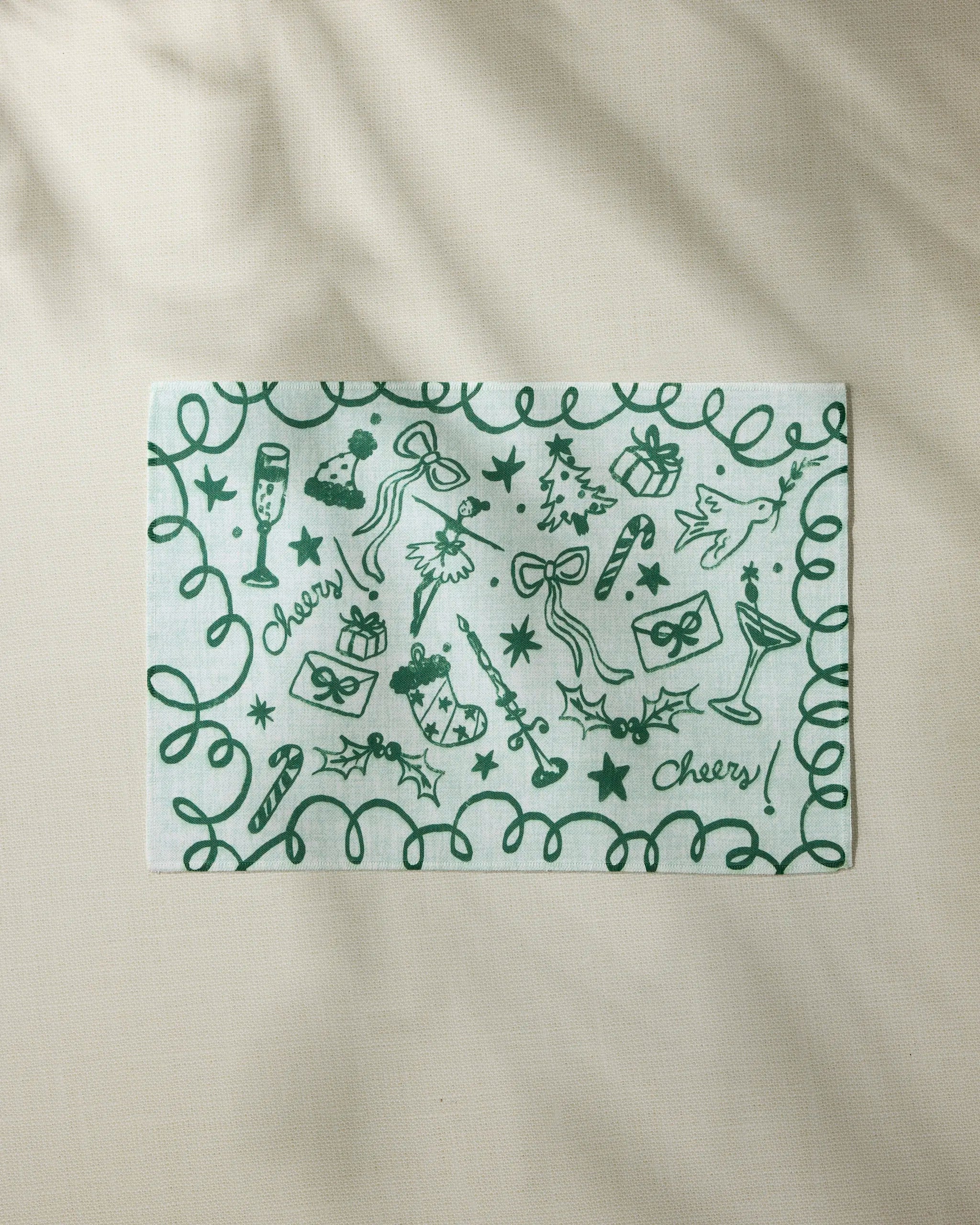 The Geometry "Doodles for Holiday" napkin features festive green holiday doodles—gifts, holly, bows, stars, champagne glasses, a Christmas tree, candy cane, and "Cheers!"—and pairs perfectly with your favorite placemats.