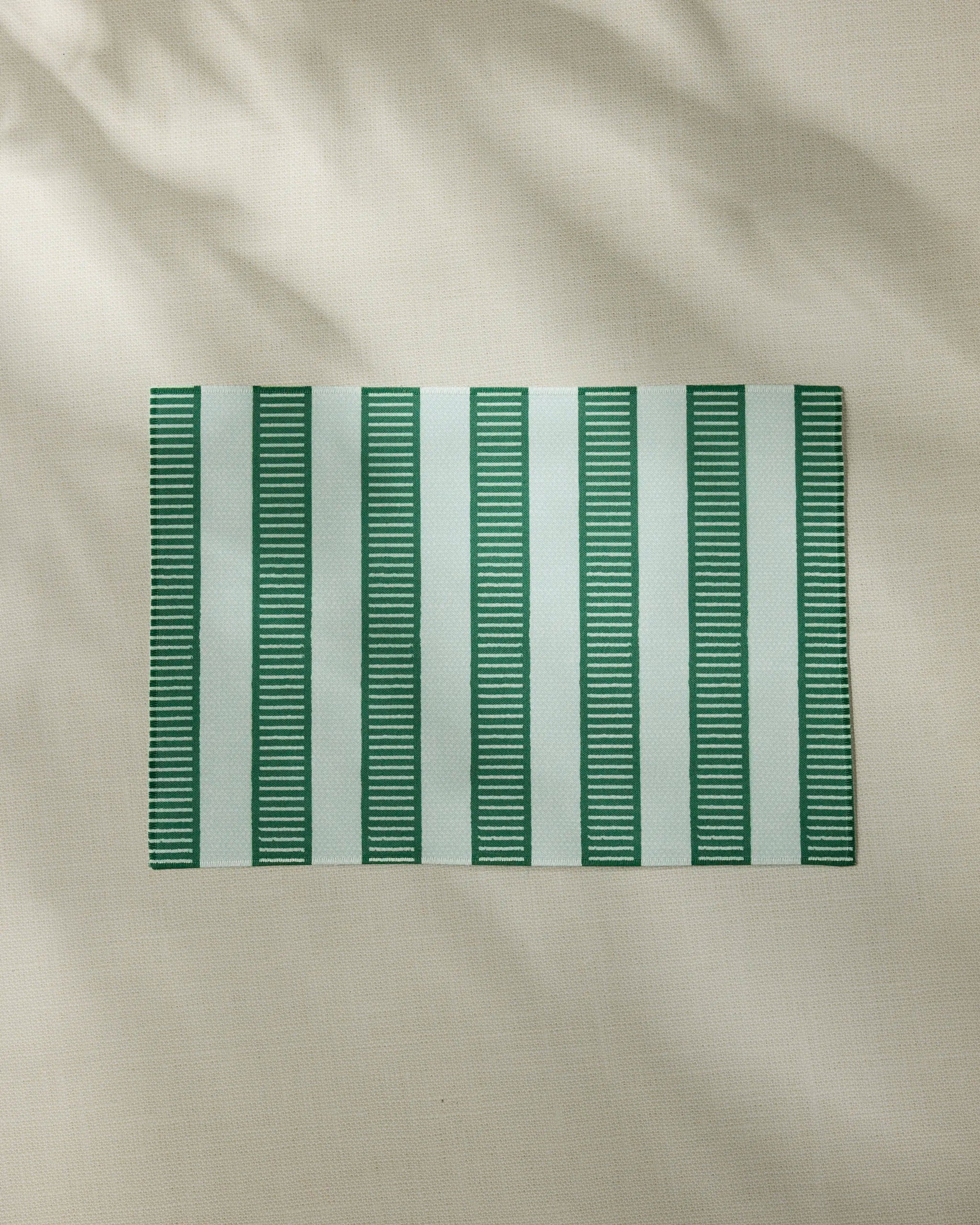 The Geometry Firstripe placemat, featuring green and white vertical stripes, rests on a light beige textured surface. Soft shadows highlight this stylish, machine-washable placemat from Geometry.