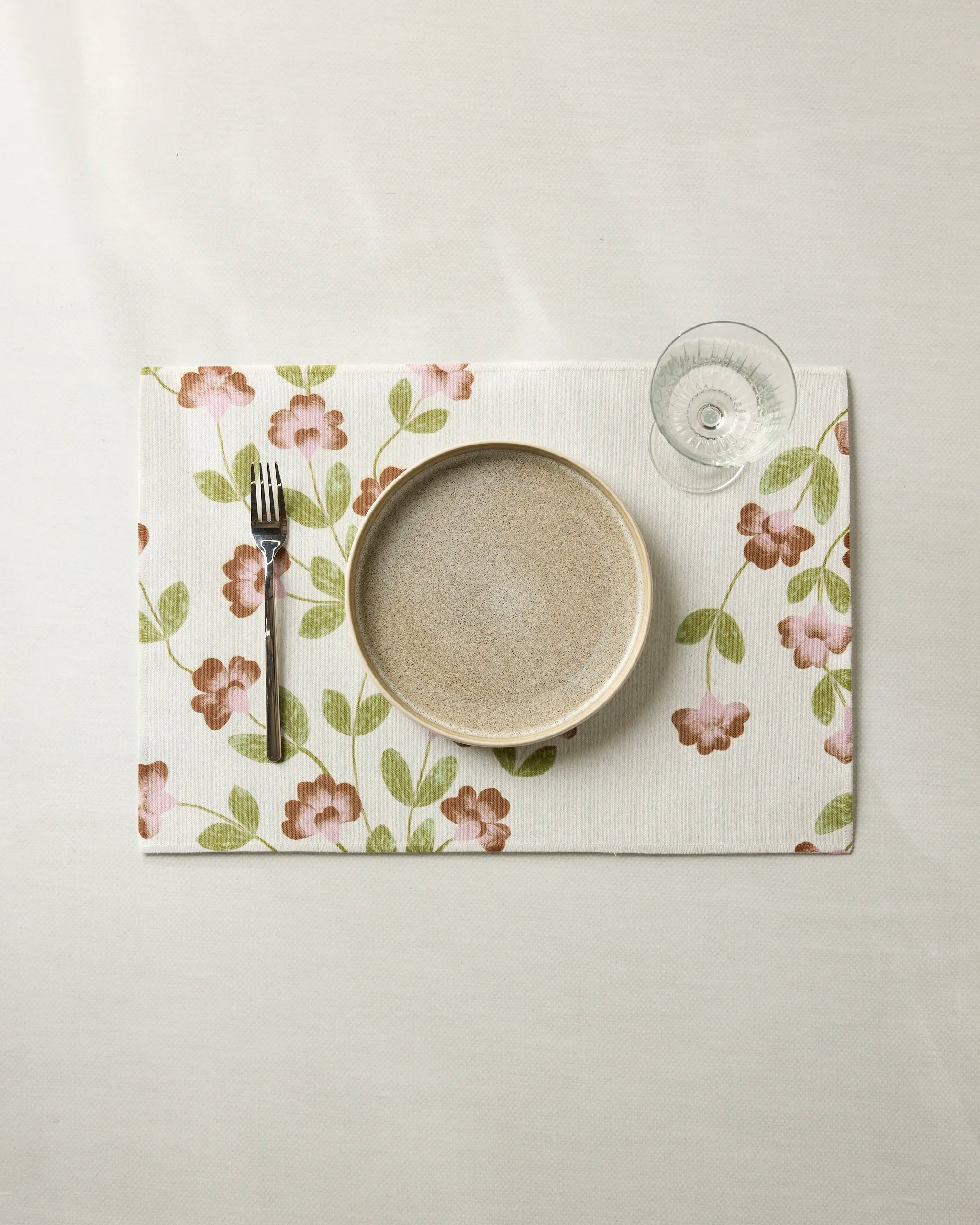 A beige plate, fork, and empty glass sit on Geometry's Flower Tower recycled placemats with pink flowers and green leaves, atop a white tablecloth.
