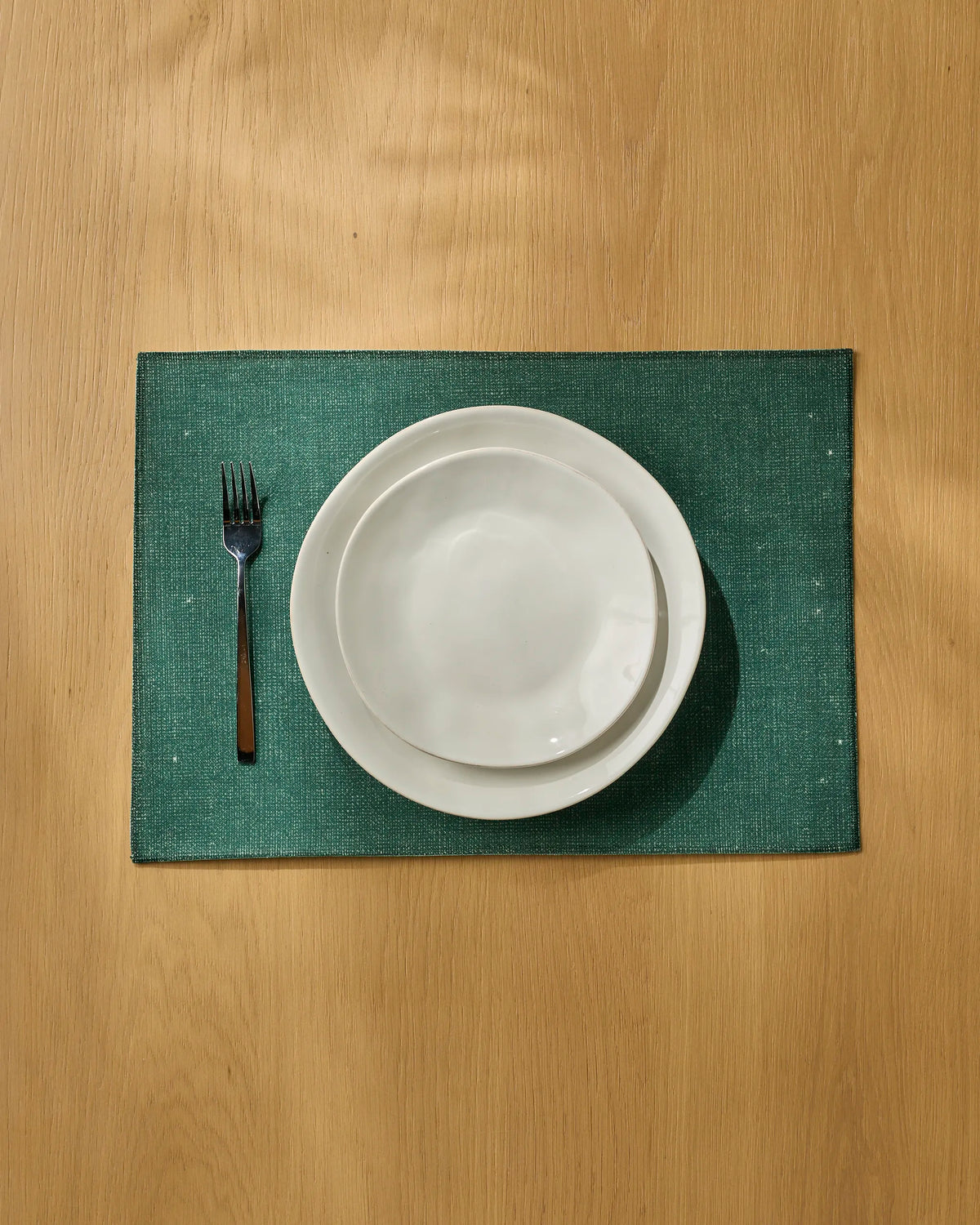 A Geometry Gather fork, bowl, and plate are neatly arranged on a green recycled placemat atop a wooden table under natural light.