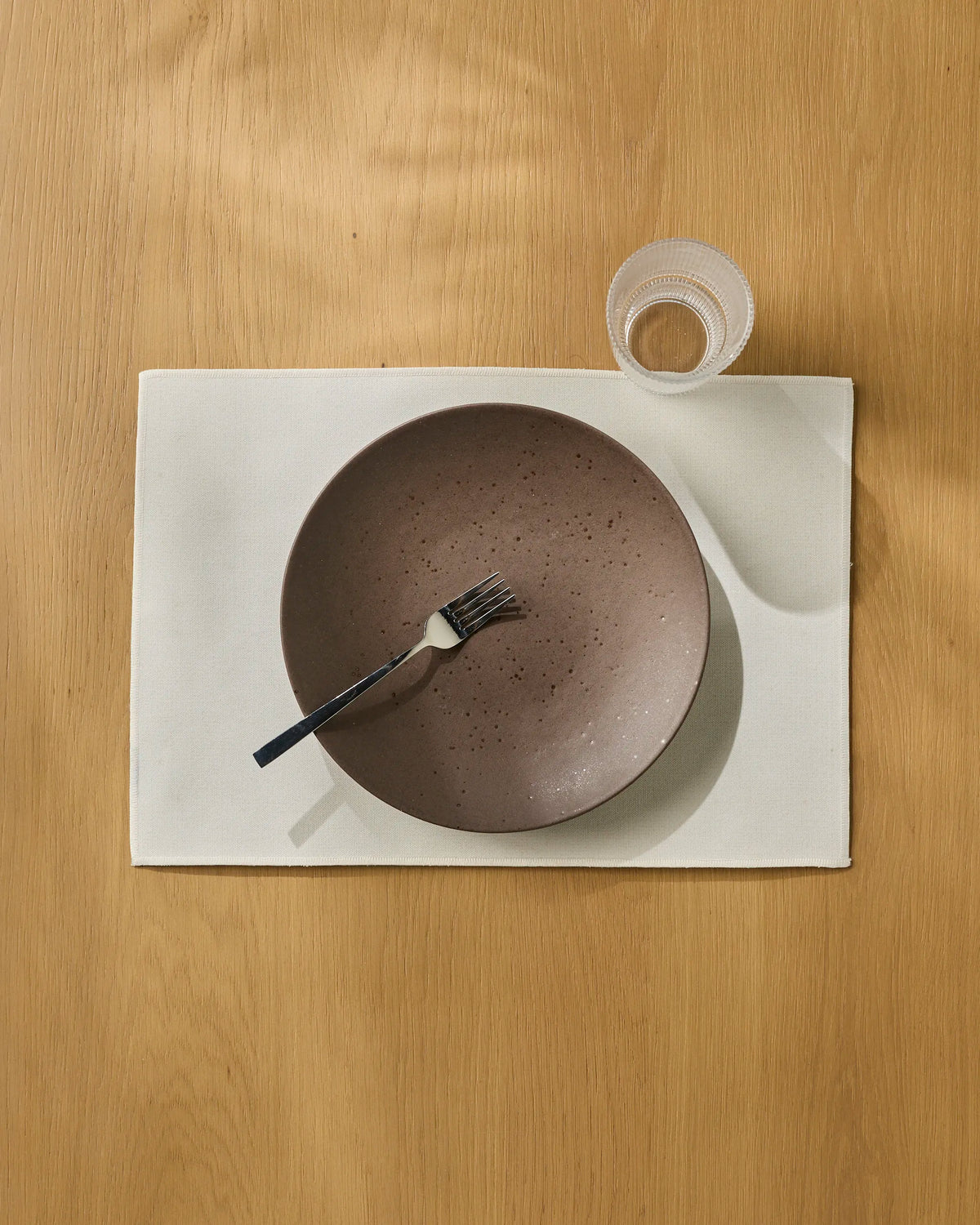 A pink speckled bowl with a fork inside rests on the white, machine-washable Geometry Gather placemat, next to a clear drinking glass, all set on a wooden table.