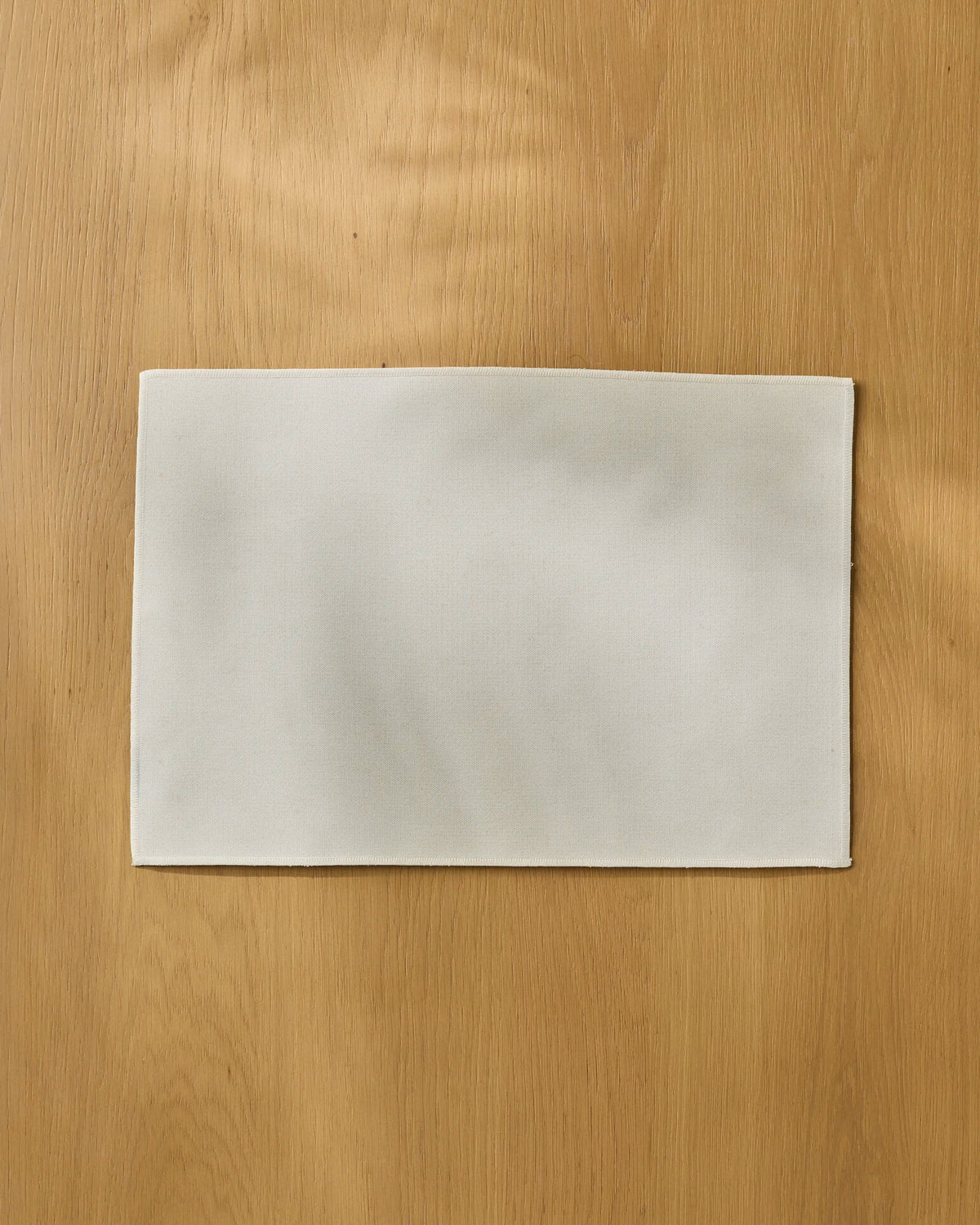 A rectangular white Gather cloth napkin by Geometry lies flat on a light wood surface, centered in the image and perfectly complementing eco-friendly placemats for a sustainable table setting.