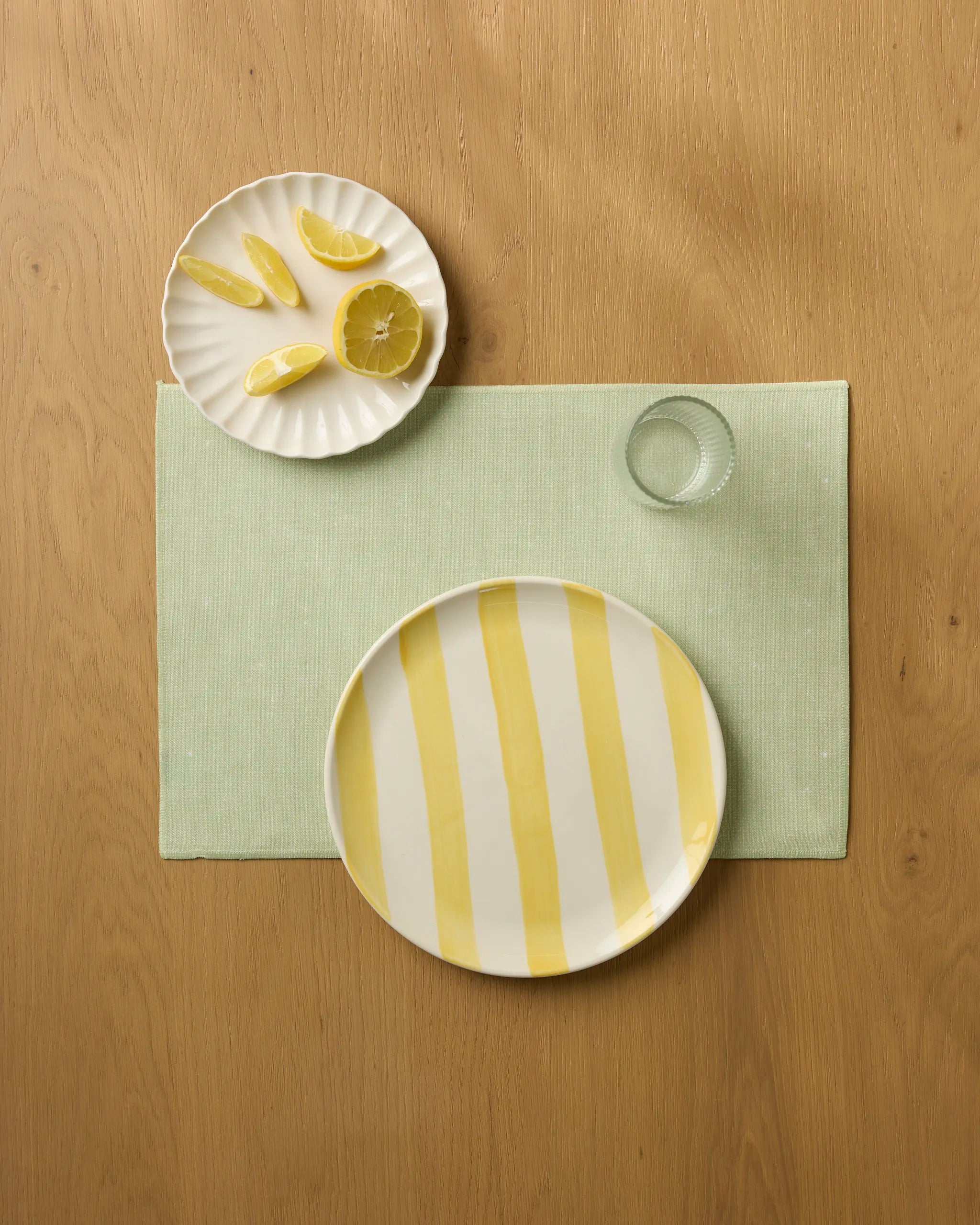 A top-down view of a table setting shows Geometry’s Greenhouse placemat in mint green, a yellow and white striped plate, a clear glass, and a small plate with lemon slices on a wooden surface.