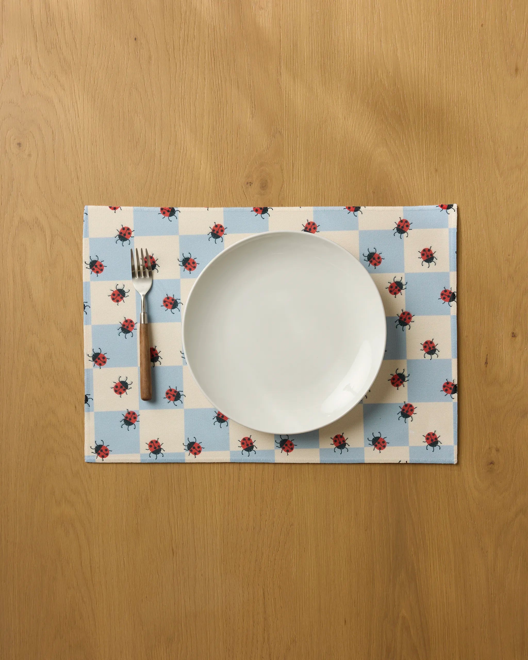 A white plate and fork sit on the Geometry Mariquita Pop placemat, featuring blue-and-white checks with red flower patterns, atop a light wooden table.