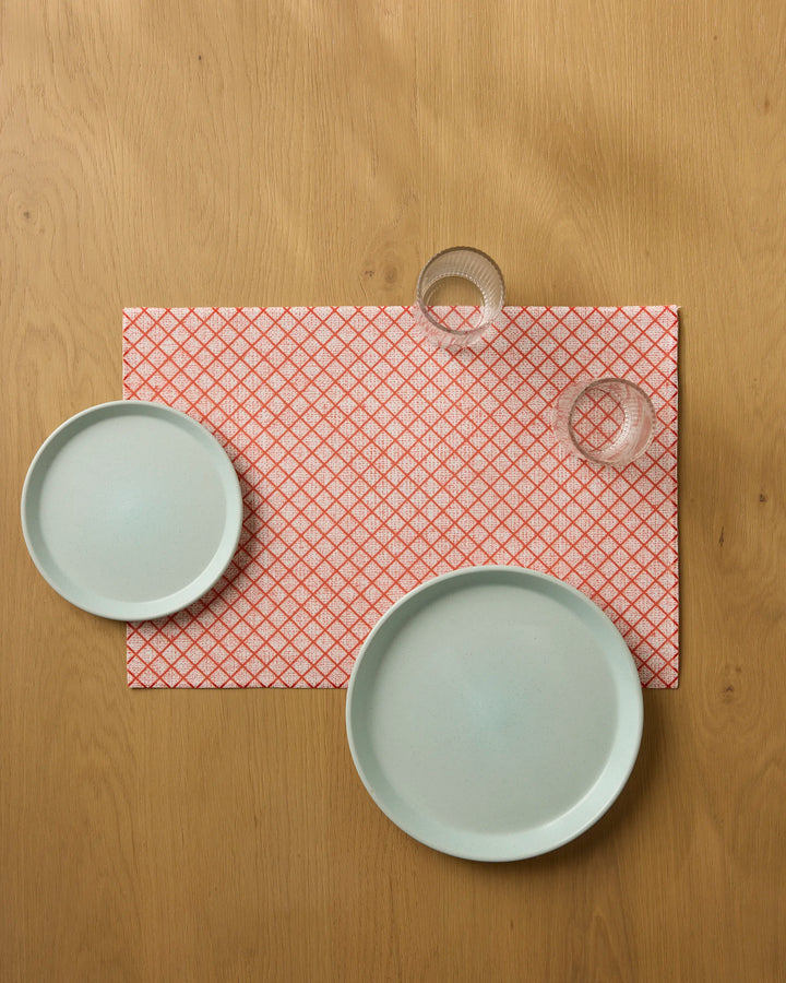 Two light blue plates and two clear glasses sit on a red-and-white patterned Market Check placemat by Geometry, placed atop a wooden table and viewed from above.