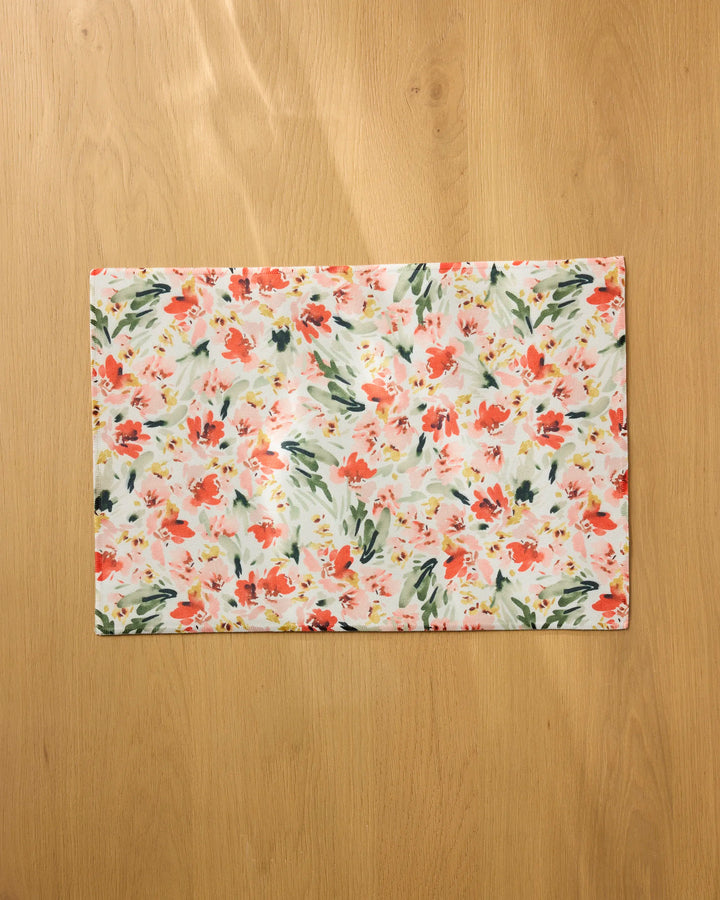 The Melissa Johnson "Painted Garden" placemat, featuring a floral pattern in pink, red, green, and yellow, sits on a light brown wooden surface. It's rectangular and machine washable.