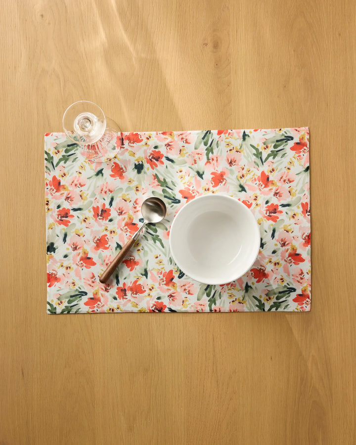 The "Painted Garden" placemat by Melissa Johnson, featuring a floral design, rests on a wooden table with a white bowl, spoon, and an empty stemmed glass. Sunlight casts gentle shadows over this serene setting.