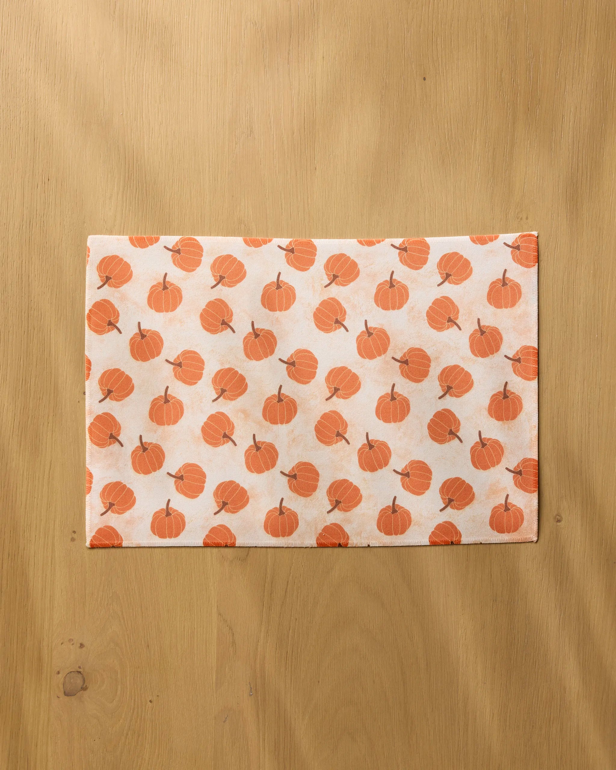 The Geometry Pumpkin Polka is a rectangular white placemat with an orange pumpkin pattern, lying flat on light wood. It's machine washable for easy cleaning and adds stylish flair to your table setting.