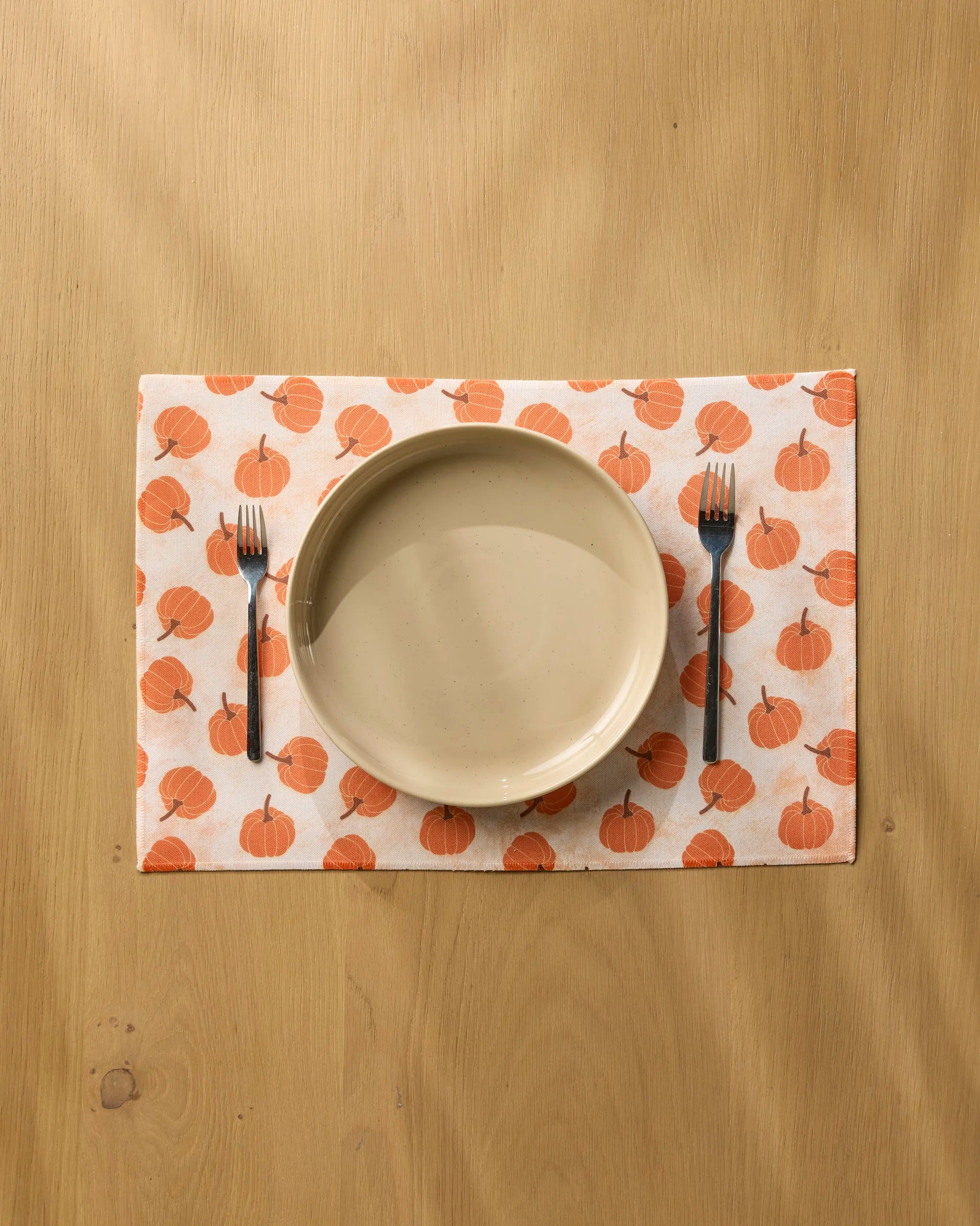 A beige plate sits on a Geometry Pumpkin Polka placemat, made from recycled materials and decorated with orange pumpkins, with a fork on each side, all placed on a light wooden table.