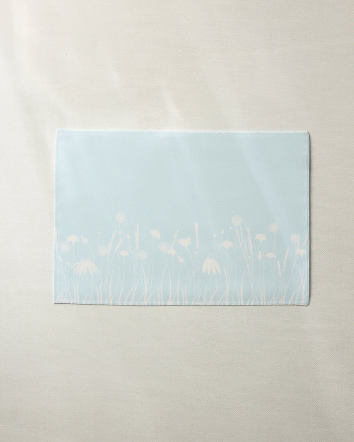 The Geometry Quiet Garden placemat features a light blue rectangular design with white floral silhouettes along the bottom edge, is machine washable, and is shown on a light beige fabric surface.