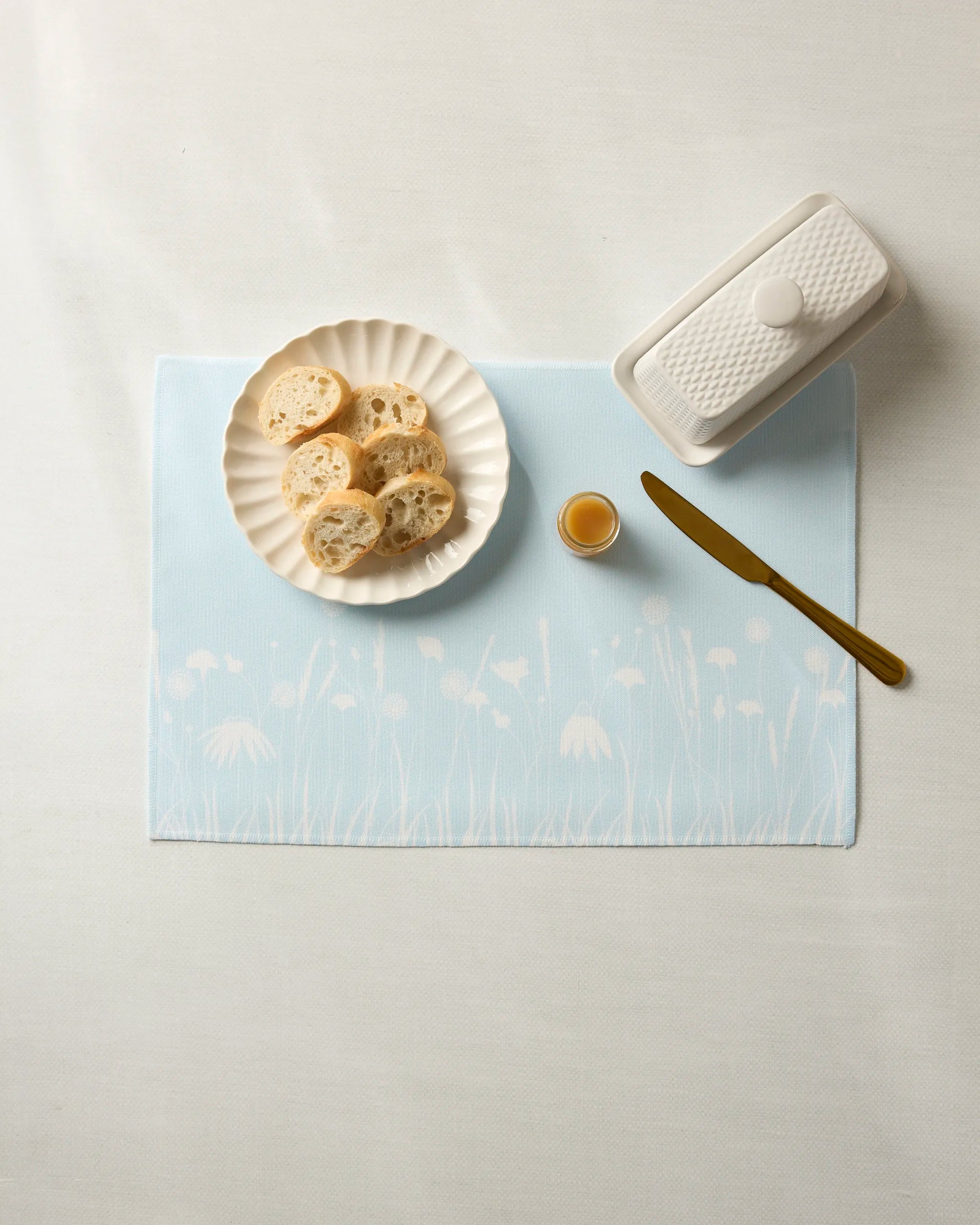 A white plate with bread, butter dish, honey jar, and gold knife sit on Geometry’s Quiet Garden machine-washable placemats featuring a blue floral pattern, all atop a white tablecloth.