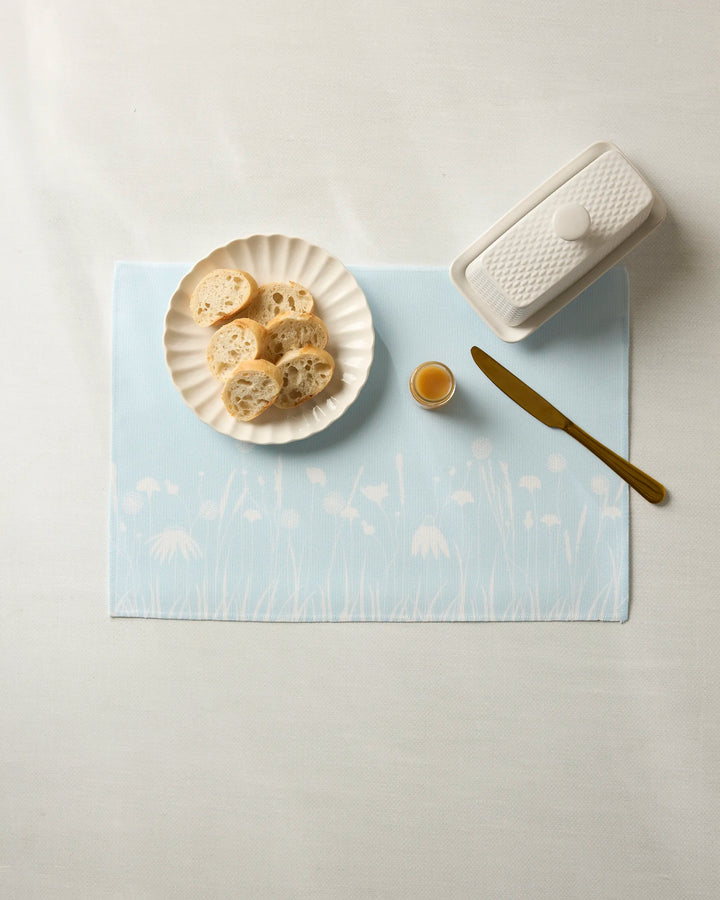 A white plate with bread, butter dish, honey jar, and gold knife sit on Geometry’s Quiet Garden machine-washable placemats featuring a blue floral pattern, all atop a white tablecloth.