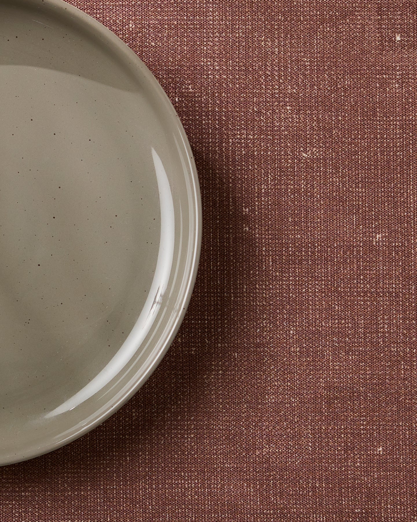 A close-up of Geometry’s eco-friendly Russet placemats showcases their style on a textured, reddish-brown background beside a light gray ceramic plate, which is partially visible and empty on the left.