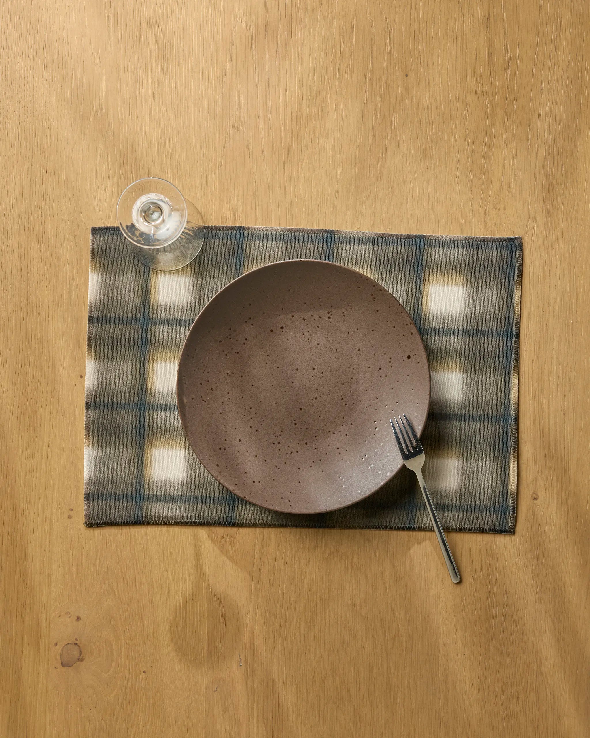 A ceramic plate and a fork rest on the eco-friendly Geometry "Toasted" placemat in blue, gray, and white hues, beside an empty wine glass atop a light wooden table.