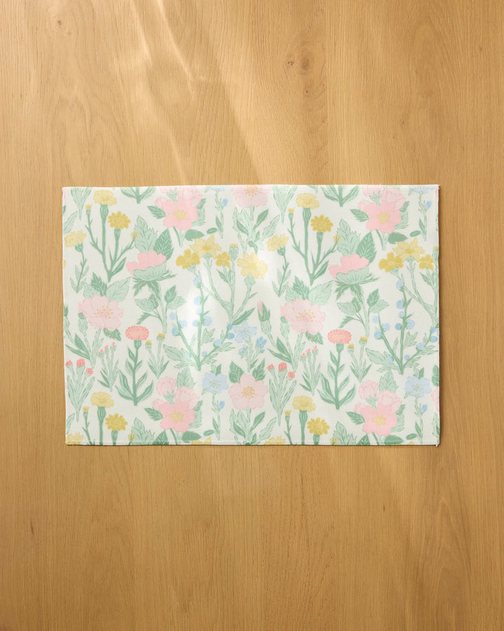 The Wildflowers in Bloom placemat by Jasmine Blooms Designs features a pastel floral pattern with pink, yellow, and blue flowers plus green leaves, shown here on a light wooden surface.