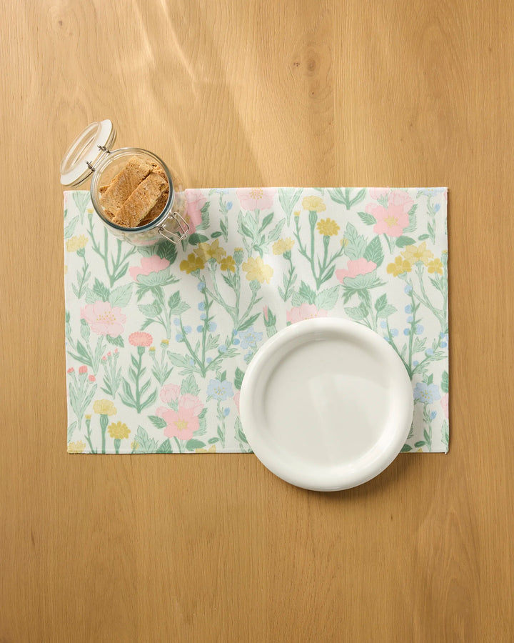 The "Wildflowers in Bloom" placemat by Jasmine Blooms Designs sits on a wooden table, holding a white bowl and a glass jar with a hinged lid filled with cookies.