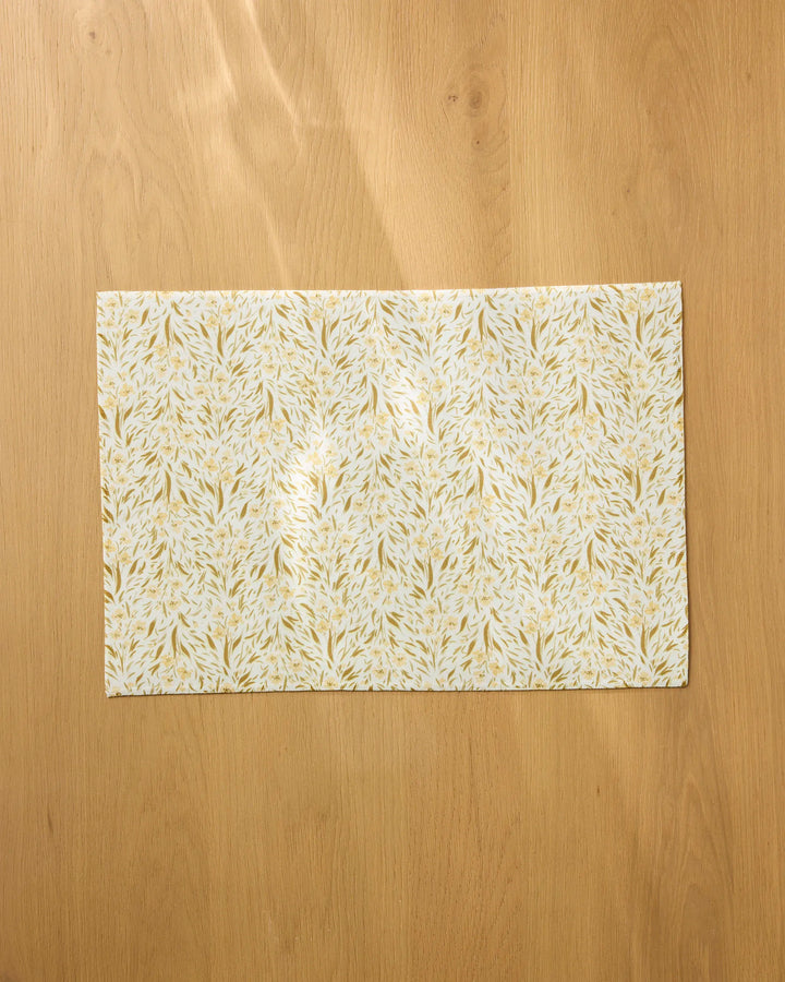 A rectangular sheet from Melissa Johnson’s Yellow Floral collection, featuring a gold and white leafy pattern, rests on a light wooden surface, evoking the look of sophisticated and durable table placemats.