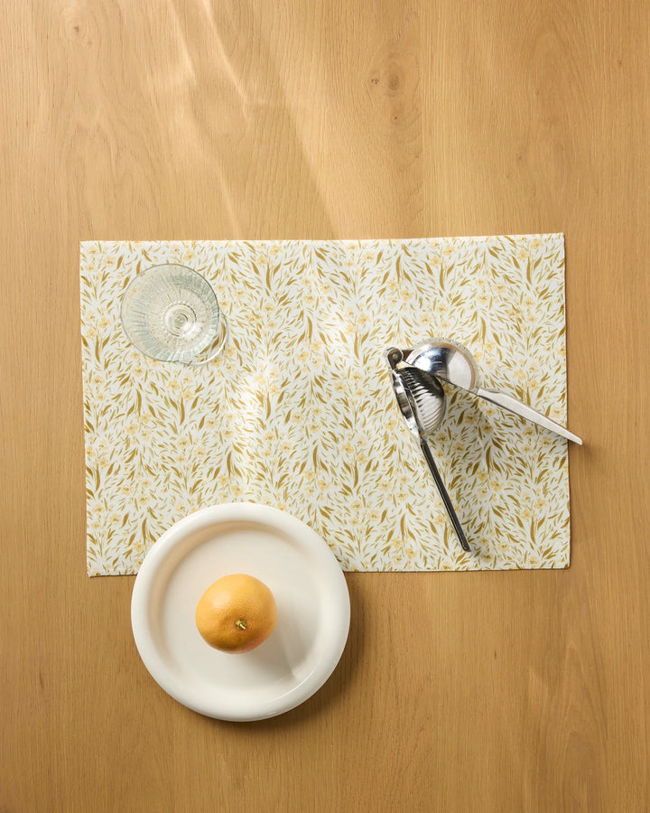 A glass, citrus squeezer, and white plate with a grapefruit sit on Melissa Johnson’s Yellow Floral placemats atop a wooden table, viewed from above.