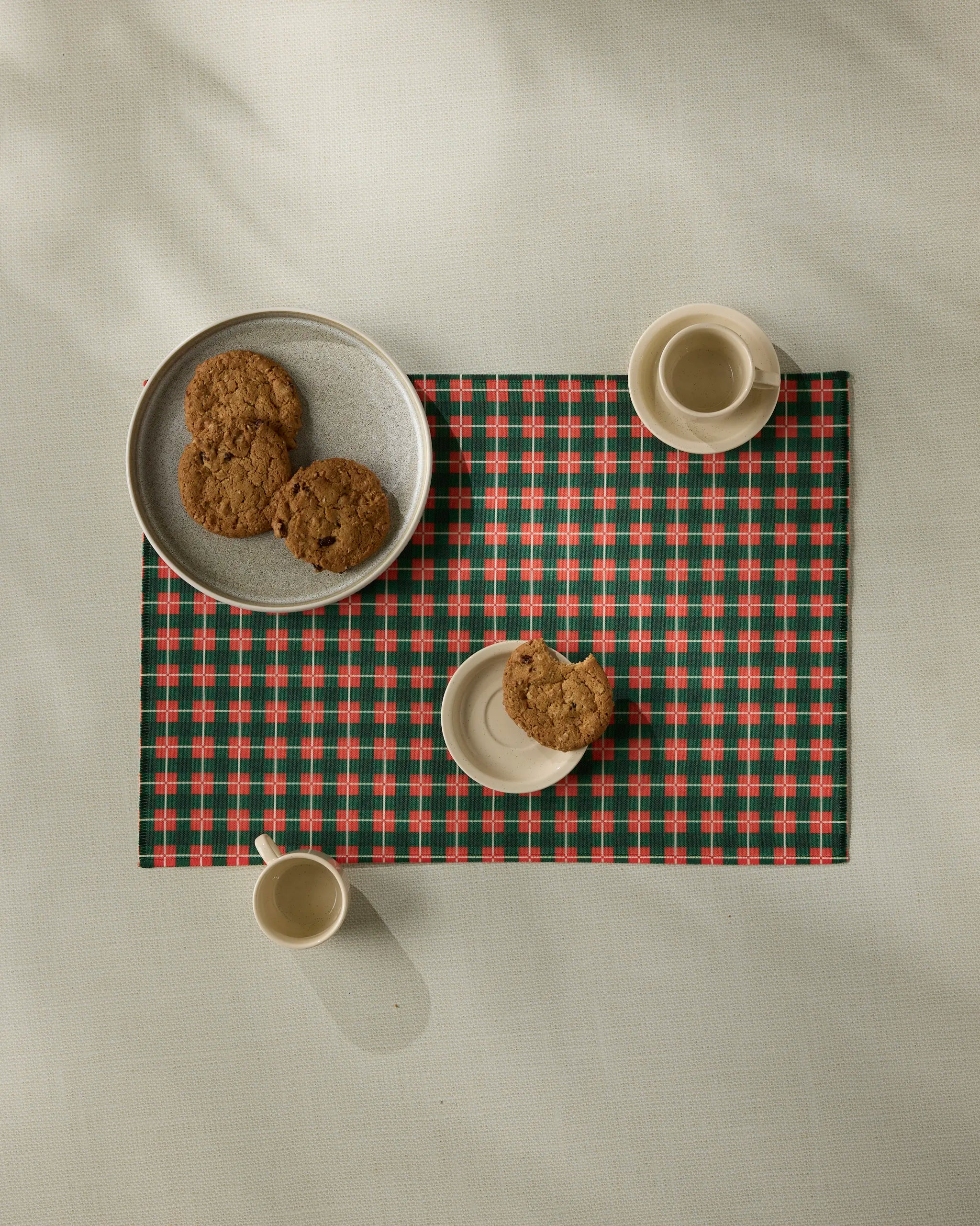 A Yuletide Green Plaid placemat by Geometry displays a small plate with a cookie, two cups of coffee, and a larger plate holding three cookies, all viewed from above on a subtly textured background.