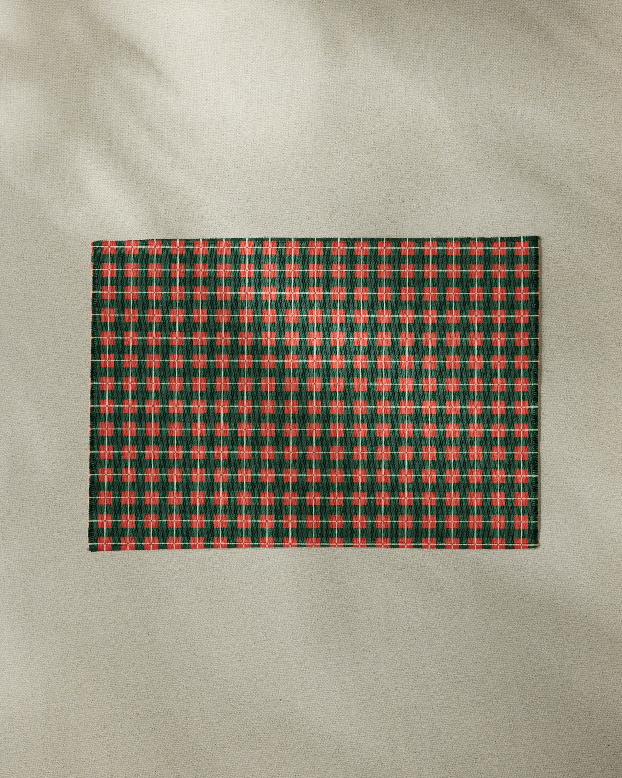 The Yuletide Green Plaid placemat by Geometry, featuring a red, green, and white plaid design, rests on a light beige textured surface with soft shadows in the background.