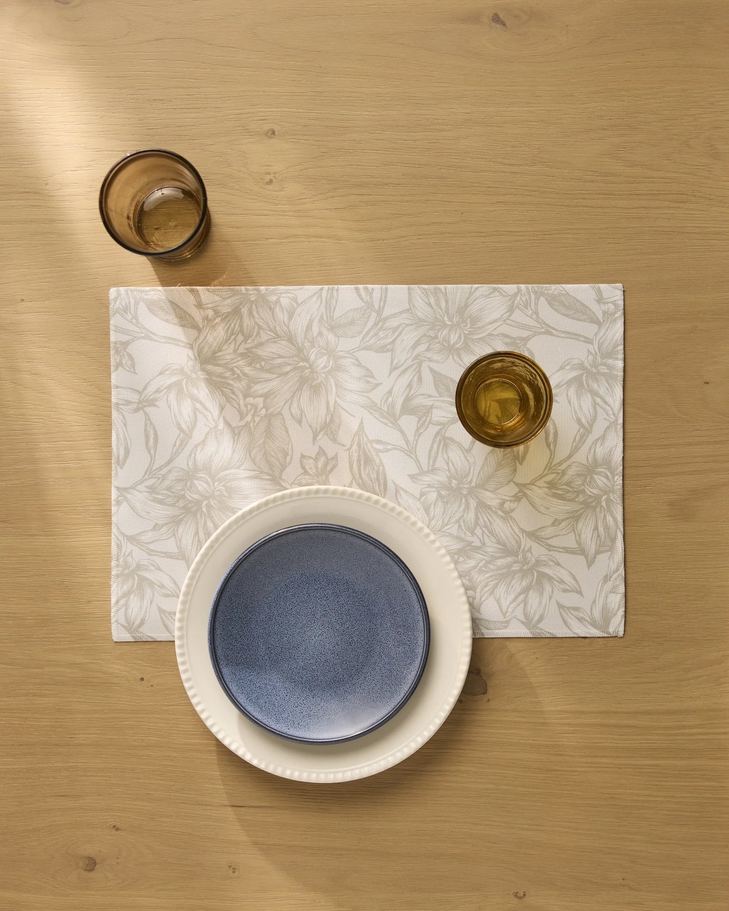 A table setting features Geometry’s Alba machine-washable placemats with a floral design, a white plate with a blue bowl, and two amber glasses on light wood, sunlight softly illuminating the scene.