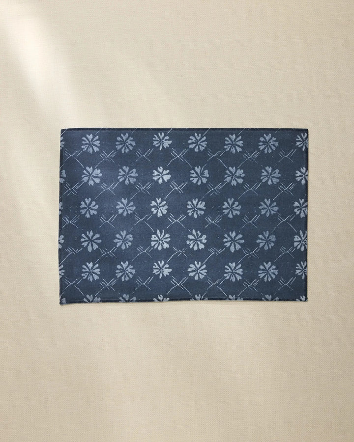 The Geometry Bonnie Indigo rectangular placemat, featuring a dark blue color and subtle light blue floral pattern, rests on a beige textured surface.
