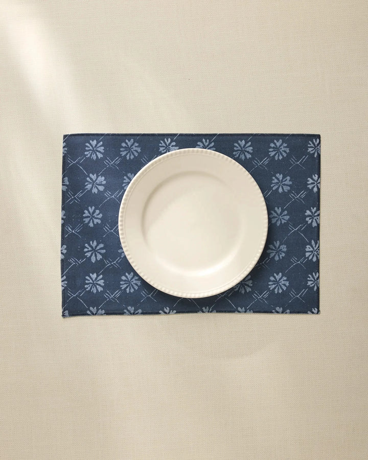 A white ceramic plate is centered on the Geometry Bonnie Indigo machine washable placemat, featuring a light floral pattern, atop a cream-colored tablecloth.