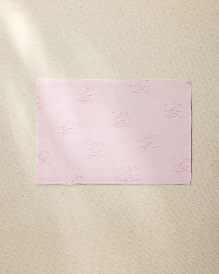 The Bubble Ditzy Dusty Rose placemat by Geometry is a rectangular pale pink cloth with subtle light purple star-like patterns, shown laid flat on a beige textured surface with soft lighting and a gentle left shadow.