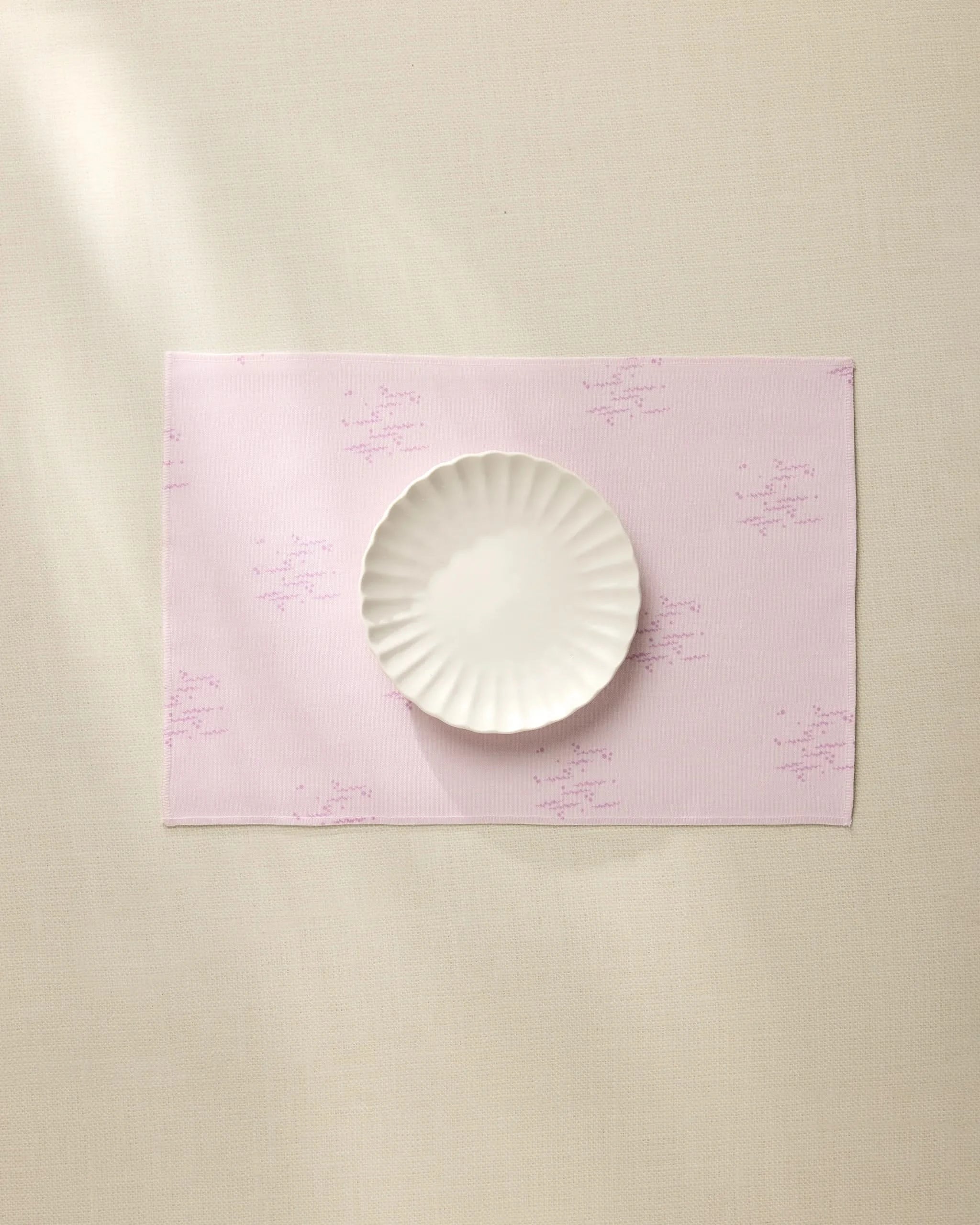 The Geometry Bubble Ditzy Dusty Rose placemat, featuring subtle lavender patterns, sits on a beige surface under soft lighting with a white scalloped-edge plate centered on top. Machine washable for easy care.