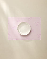 The Geometry Bubble Ditzy Dusty Rose placemat, featuring subtle lavender patterns, sits on a beige surface under soft lighting with a white scalloped-edge plate centered on top. Machine washable for easy care.