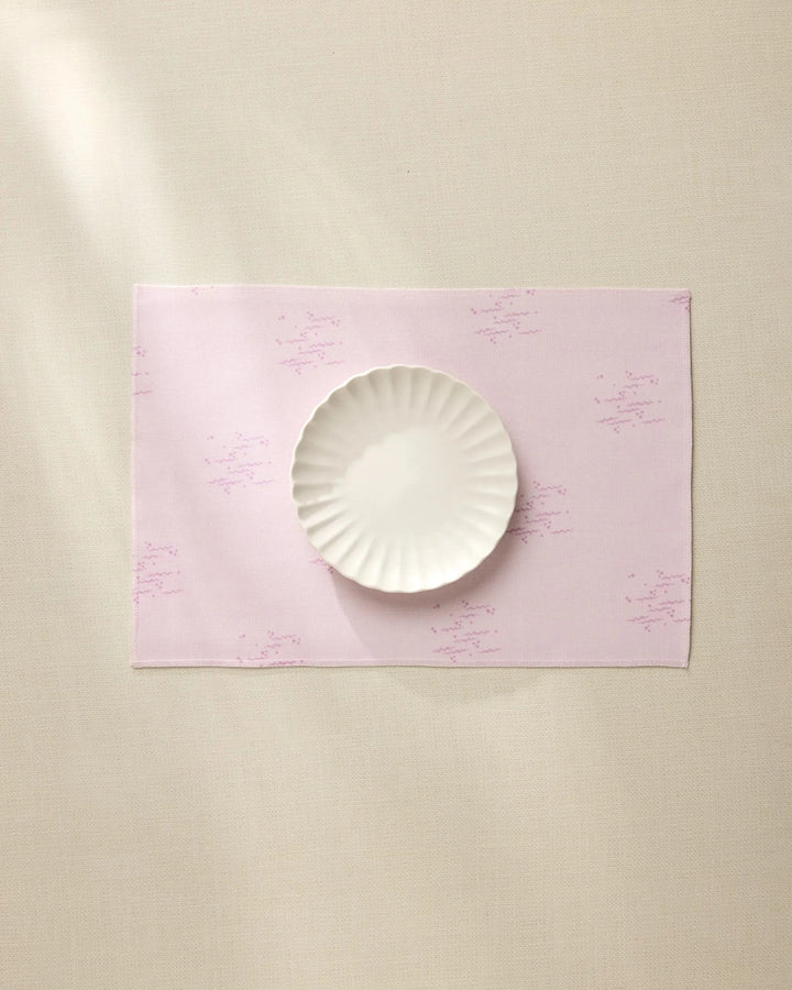 The Geometry Bubble Ditzy Dusty Rose placemat, featuring subtle lavender patterns, sits on a beige surface under soft lighting with a white scalloped-edge plate centered on top. Machine washable for easy care.
