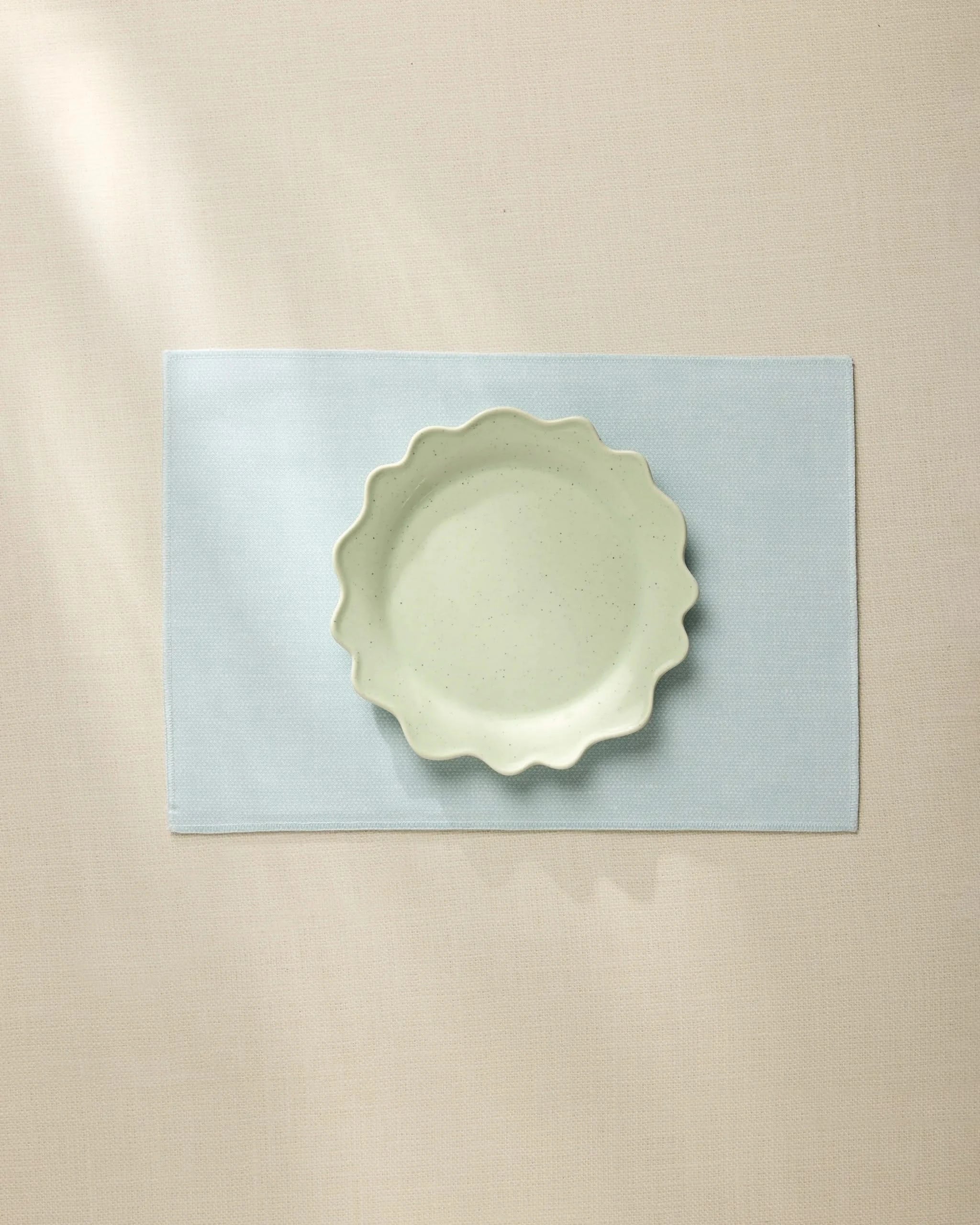 A light green, wavy-edged ceramic bowl sits centered on the Geometry Chambray Light Blue placemat, which rests on a beige tablecloth under soft, natural light.