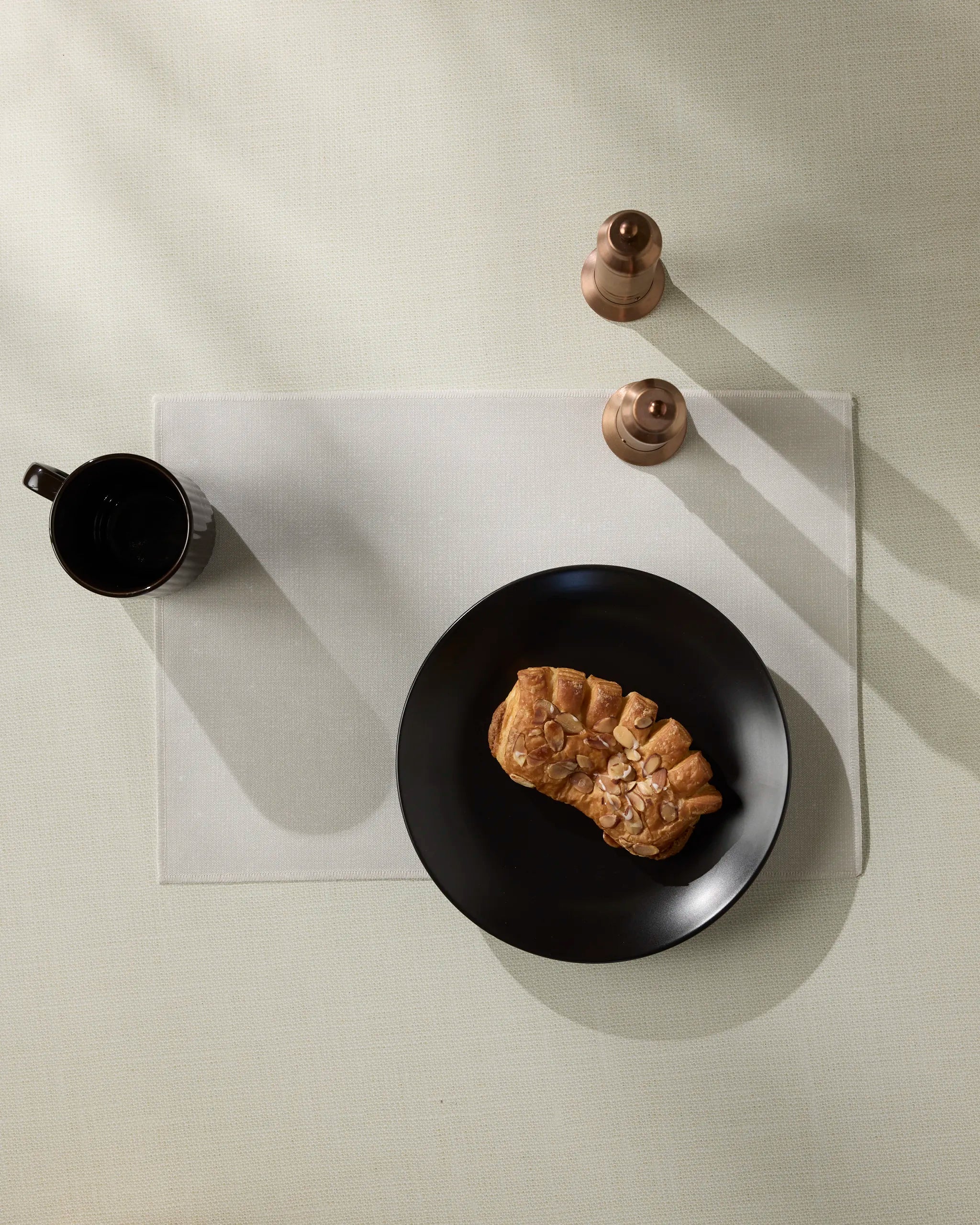 A black Geometry Frost plate with a pastry rests on an eco-friendly placemat, accompanied by a matching mug and two copper salt and pepper shakers. Bright light highlights the sleek setup on the pale table.