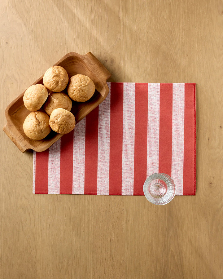 A wooden tray with six bread rolls is on a light wood table beside the Kailee Wright Peppermint Stripe placemat in red and white, which is machine washable, with a clear glass resting on it.