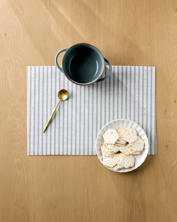 A dark gray bowl, gold spoon, and white plate with square crackers are arranged neatly on Kailee Wright's Stone Stripe placemats atop a wooden table.